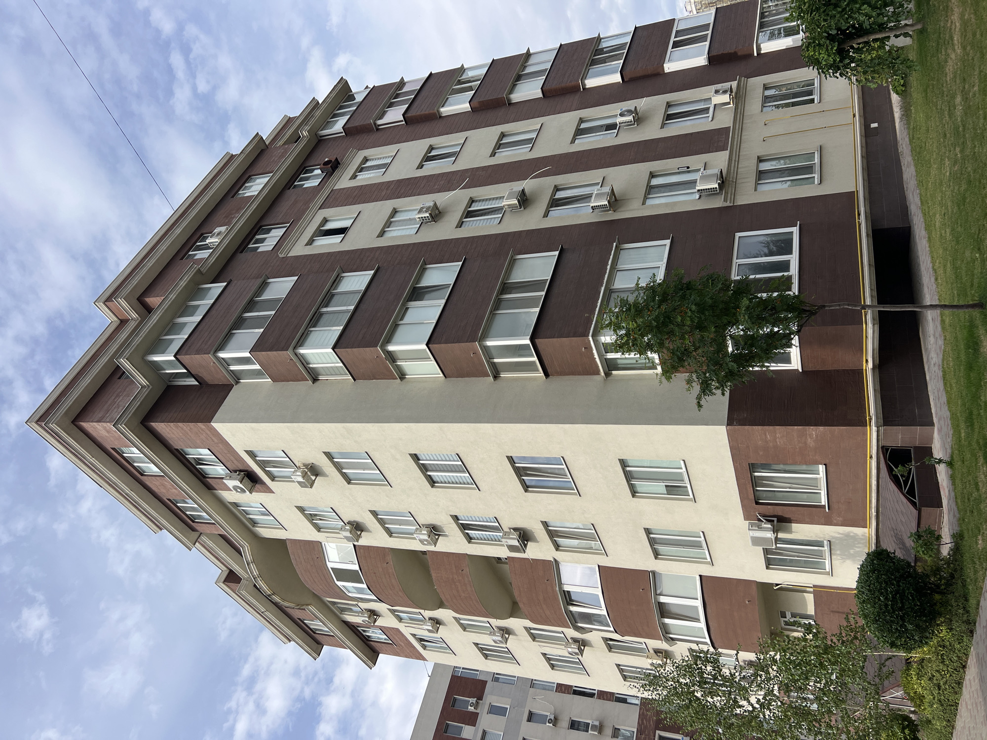 The image shows a tall, multi-story apartment building located in Kryzhanivka, Ukraine. The building has a distinctive facade with alternating sections of brown and beige siding. There are many windows visible on the building, and several air conditioning units are mounted on the exterior walls. The building is surrounded by green grass and some trees, and the sky above is partly cloudy. In the background, another apartment building is partially visible. There are no people or vehicles in the image. The scene appears to be a daytime shot.