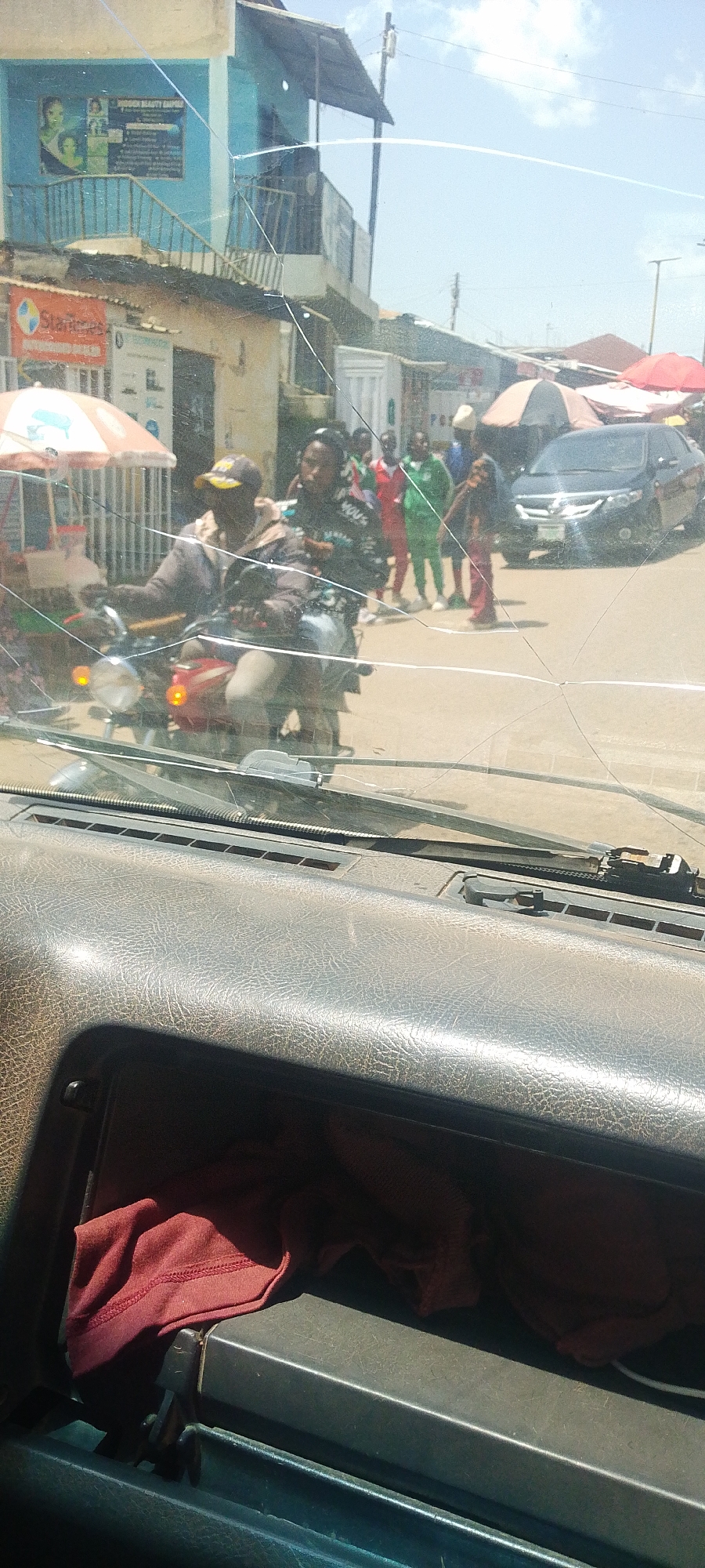 A cracked windshield obscures a view of a busy street scene in Jos, Nigeria. In the foreground, a dark grey dashboard is visible, with a folded red cloth in a compartment. In the middle ground, a man wearing a cap and a brown jacket rides a red motorcycle, with a woman on the back. Both appear to be looking ahead. Behind them, several people in athletic wear, predominantly green and red, stand on the side of the road. A black Toyota sedan is parked further down the street, and various market stalls with umbrellas are set up along the roadside. The buildings in the background are mostly painted in light colors, with one prominently blue structure featuring a poster advertising "TYSOON BEAUTY CENTER". The sky is clear and blue with scattered clouds, suggesting it is daytime, likely late morning or early afternoon. The overall atmosphere appears to be one of everyday commerce and street activity.