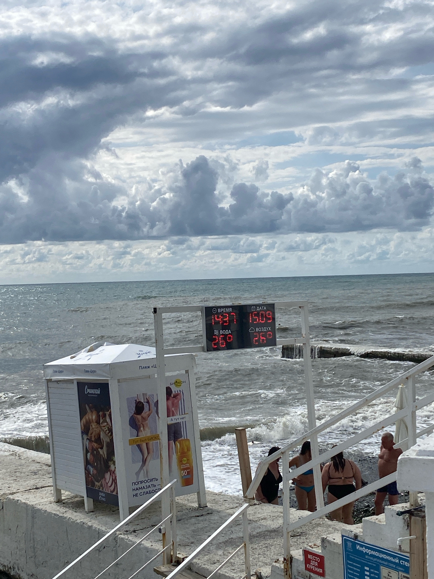 The image shows a cloudy day at a beach in Sochi, Russia. A digital display board is visible, showing the time as 14:37, the date as 15.09, the water temperature as 26 degrees Celsius, and the air temperature as 26 degrees Celsius. In the foreground, there is a small white kiosk with advertisements. A set of white stairs leads down to the beach, where several people are present. The sea is rough with waves breaking on the shore.