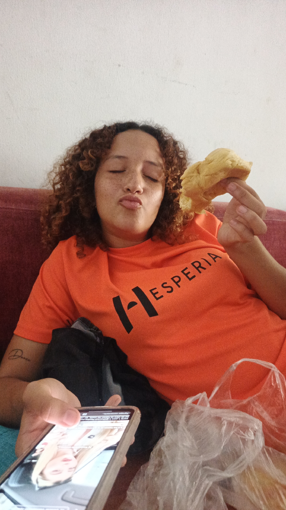 A woman with curly brown hair, freckles, and closed eyes is reclining on a red upholstered surface. She is wearing an orange t-shirt with the word "HESPERIA" printed on it in black. She is holding a piece of bread or pastry in her right hand, while holding a cell phone in her left. The cell phone screen shows a video or image of a person. There is a black bag and a clear plastic bag next to her. The setting appears to be indoors. I cannot verify the exact location as Playa El Agua, Venezuela from the current image.