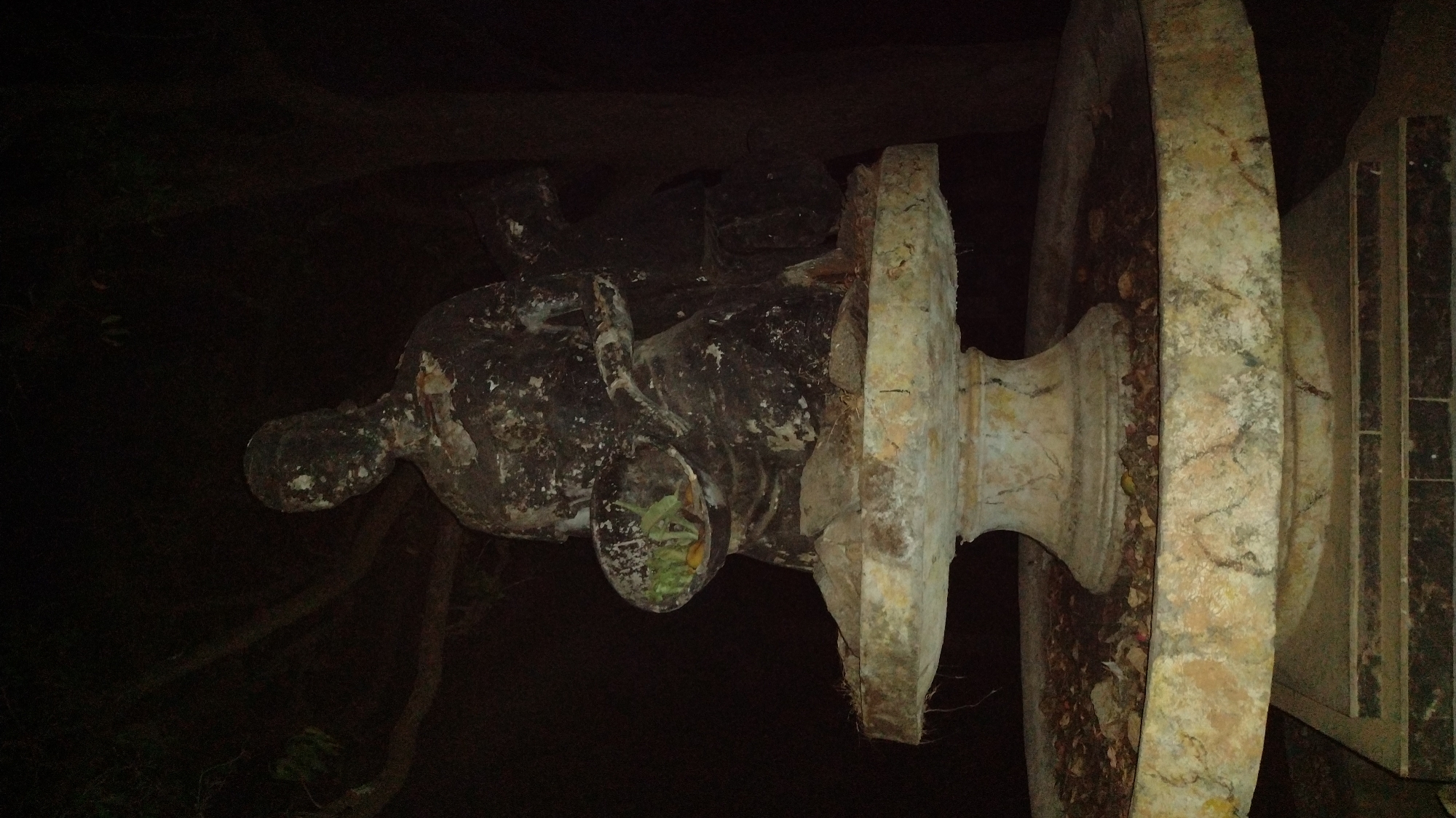 A low-angle, close-up shot shows an old, damaged statue in a dark environment. The statue is of a human-like figure with a large, circular base. The statue is heavily weathered, with chipped paint and visible cracks. There are plants growing out of one area of the statue. The background is dark, suggesting it is nighttime or a dimly lit location. While the statue might be located in Jos, Nigeria, as requested, there are no visual cues in the image to confirm this location. The image does not show people, interactions, or actions.