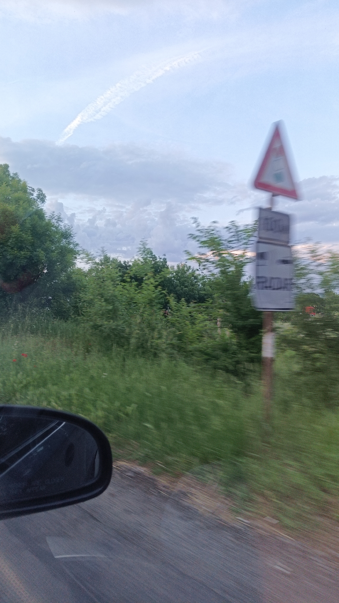 The photograph appears to be taken from inside a moving vehicle, likely a car, looking out of the side window. The road is visible at the bottom of the frame and a car side mirror is partially visible. The landscape includes roadside vegetation, predominantly green. In the background, there are trees and a cloudy sky with a contrail. A road sign is visible, consisting of a triangular warning symbol above a rectangular sign. The words on the sign are unreadable due to motion blur. The general environment suggests a rural or semi-rural setting. The location of the city of Burgas, Bulgaria, is not verifiable from the image.