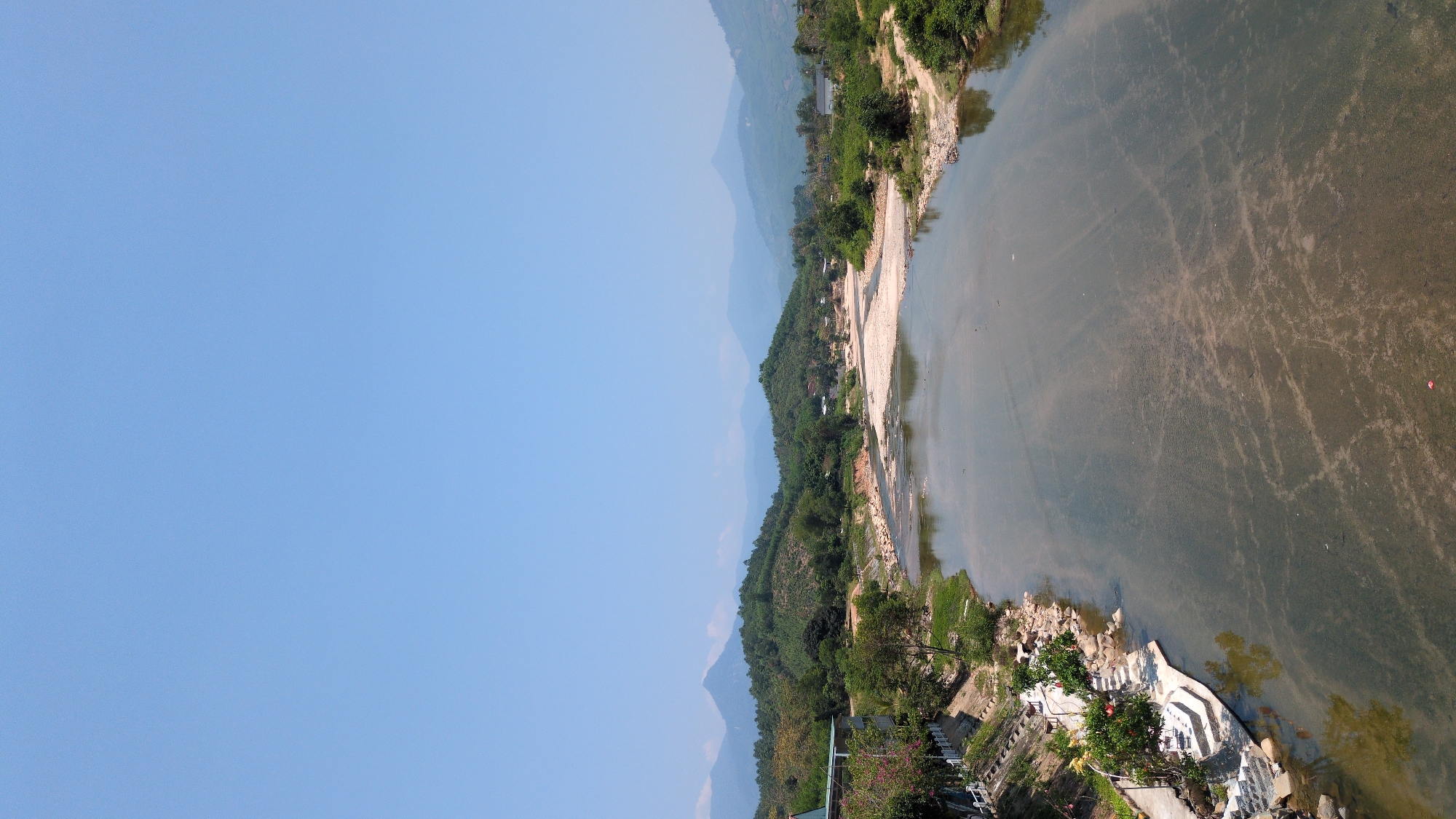 The image shows a view of the Krong Hanh river in Nam Khánh Vĩnh, Vietnam. The river is bordered by sandy banks and vegetation. Mountains are visible in the distance. The sky is clear and blue. A concrete structure is visible on the riverbank. The water is murky.