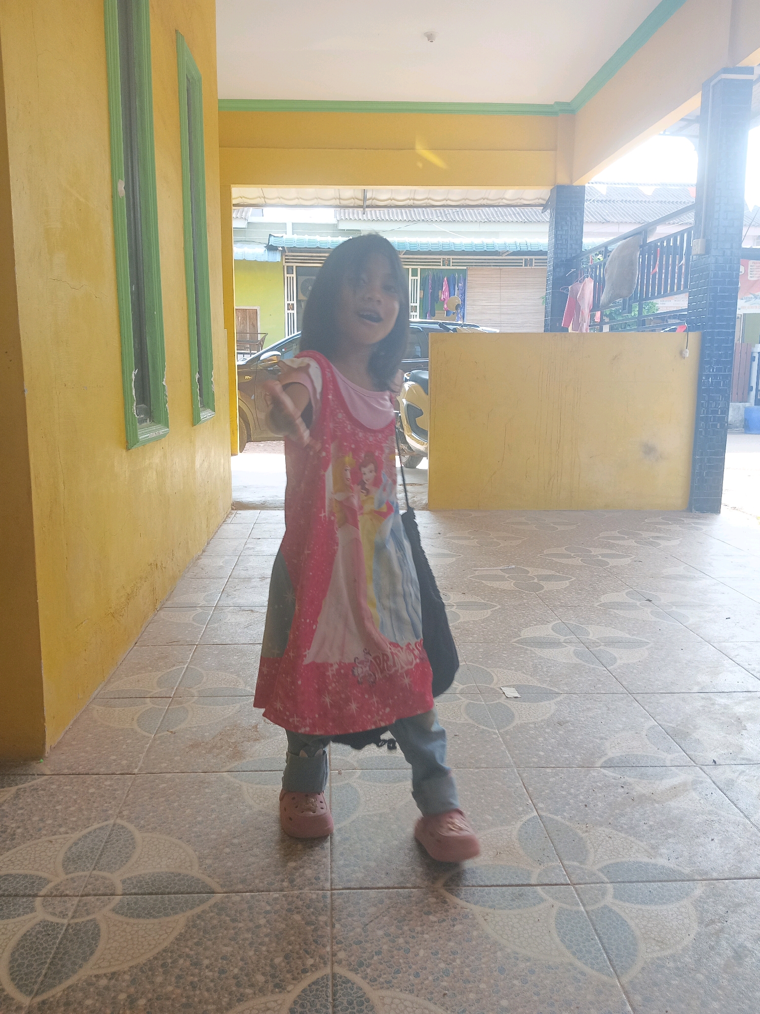 The image captures a young girl standing on a tiled porch with a yellow wall to her left and a yellow overhead structure above. She is wearing a pink dress with images of Disney princesses, blue jeans, and pink shoes. A black bag is slung over her right shoulder. The tiled floor features a repeating floral pattern in blue and gray. In the background, a car is visible, and beyond that, houses and laundry hanging on a line. The lighting suggests it is daytime, with sunlight casting shadows. The architecture, including the yellow walls and tiled porch, is common in residential areas of Batam, Indonesia.