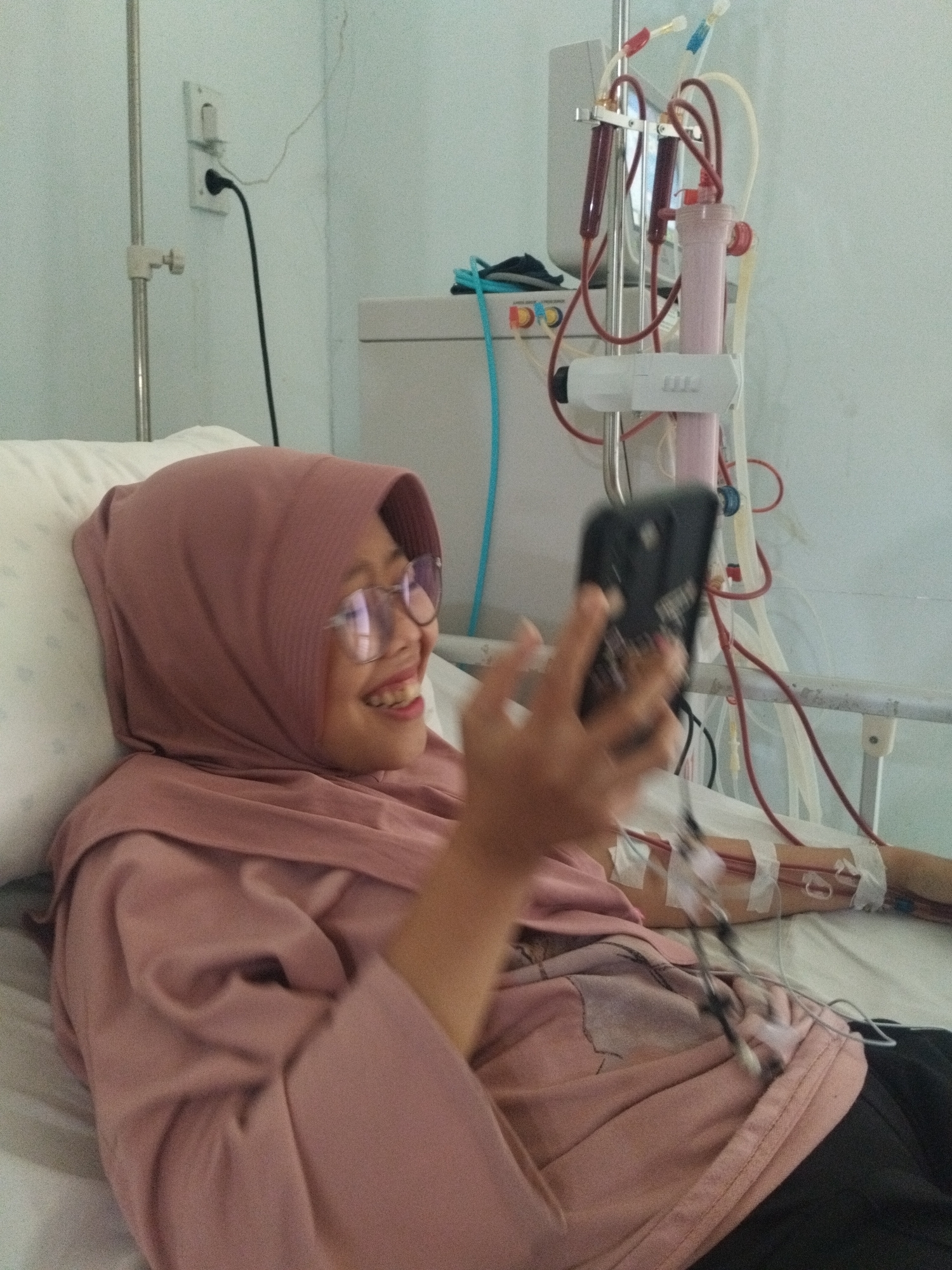 A woman in a hijab is lying in a hospital bed, smiling and holding a smartphone. Her arm is connected to medical tubing for dialysis. The location is most likely in a hospital in the city of Lhokseumawe, Indonesia. A dialysis machine is next to the bed. An electrical outlet is visible on the wall.