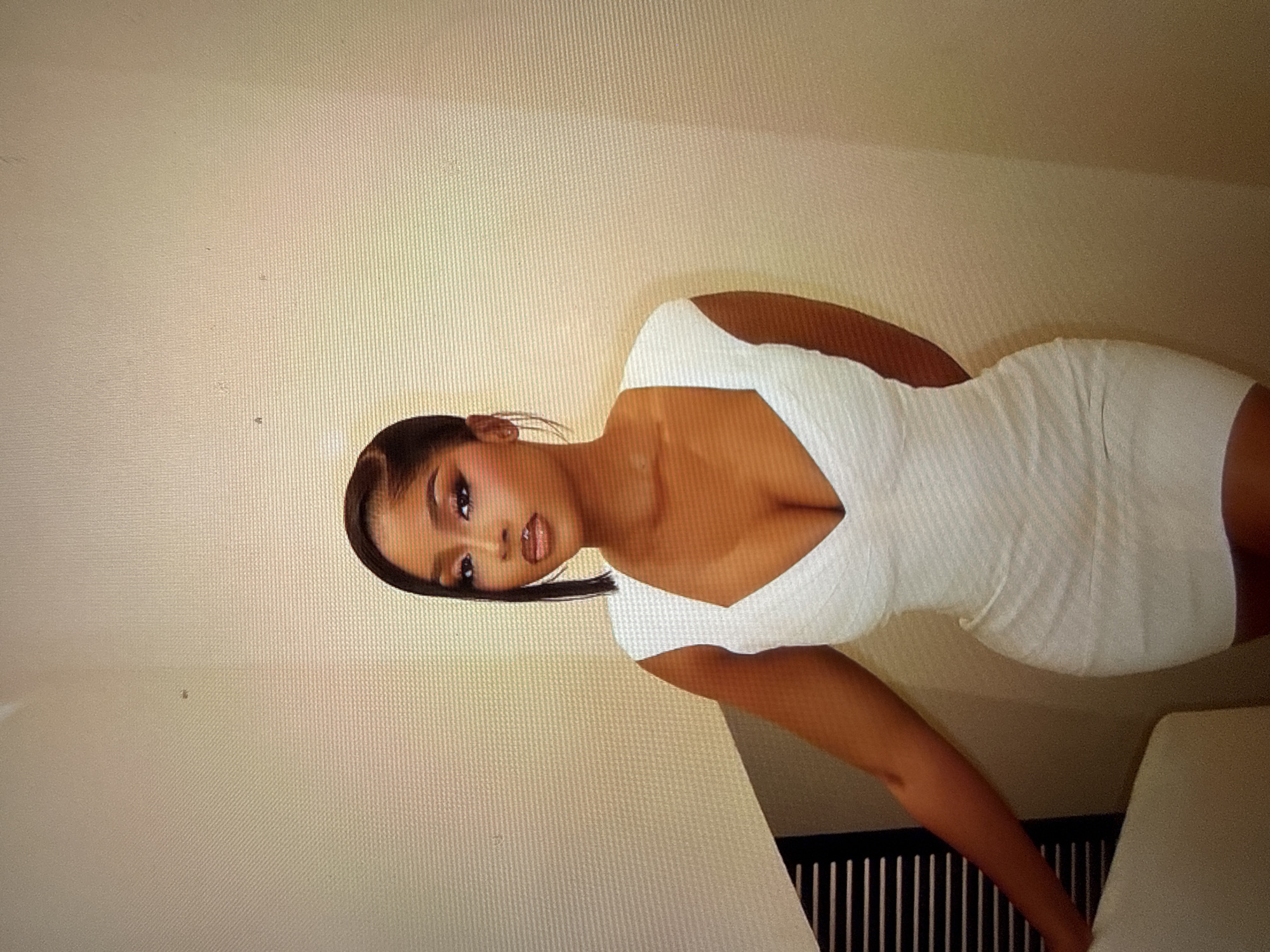 A woman wearing a white bodycon dress is positioned in the center of the frame against a textured beige wall. Her hair is pulled back with a side part, and she is wearing makeup. Her left arm is visible to the left of the frame, and her right arm is behind her back. The dress has a deep V-neckline. To the left of the woman, a dark, slatted object is partially visible, suggesting an interior setting. The lighting appears to be artificial and diffused, casting soft shadows. The overall impression is of a posed portrait. There is no identifiable information to suggest this image is located in Abuja, Nigeria.