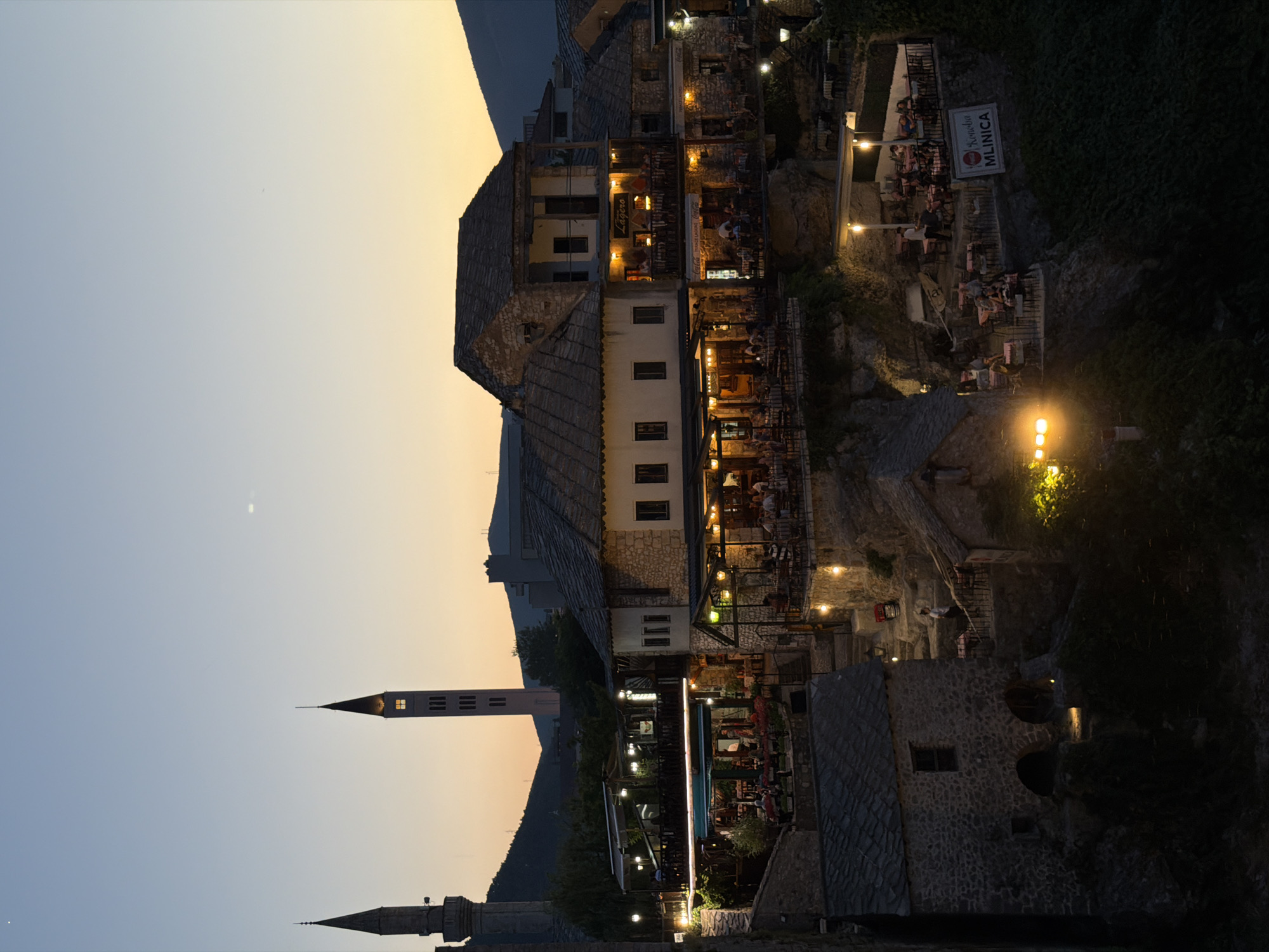 This is a twilight photograph of the Old Town of Mostar, Bosnia and Herzegovina, taken from an elevated vantage point looking down onto a series of restaurants and historic stone buildings. Two prominent minarets are visible in the background, one to the left and another taller, more slender one to the right, both silhouetted against the pale orange and blue twilight sky.
The foreground and midground are dominated by multi-level stone structures with pitched, dark-tiled roofs. These buildings appear to be part of a hillside cascading down towards a river, though the river itself is not visible. The structures are illuminated by a warm, artificial light emanating from within and from lamps strategically placed on terraces and walkways.
Several outdoor dining areas are set up on these terraces, populated by numerous patrons seated at tables. The tables are covered with white or red tablecloths, and some are adorned with flowers. Waitstaff are visible serving customers. One sign reads "konoba MLINICA" in large white letters on a red background.
The architecture is rustic and traditional, with visible stone masonry and wooden accents. Steep, stone staircases wind between the levels, also lit by lamps. The hillside is partially covered in dense green foliage, which appears as dark masses in the fading light. The overall impression is one of a lively, historic urban setting bathed in the soft glow of evening light.