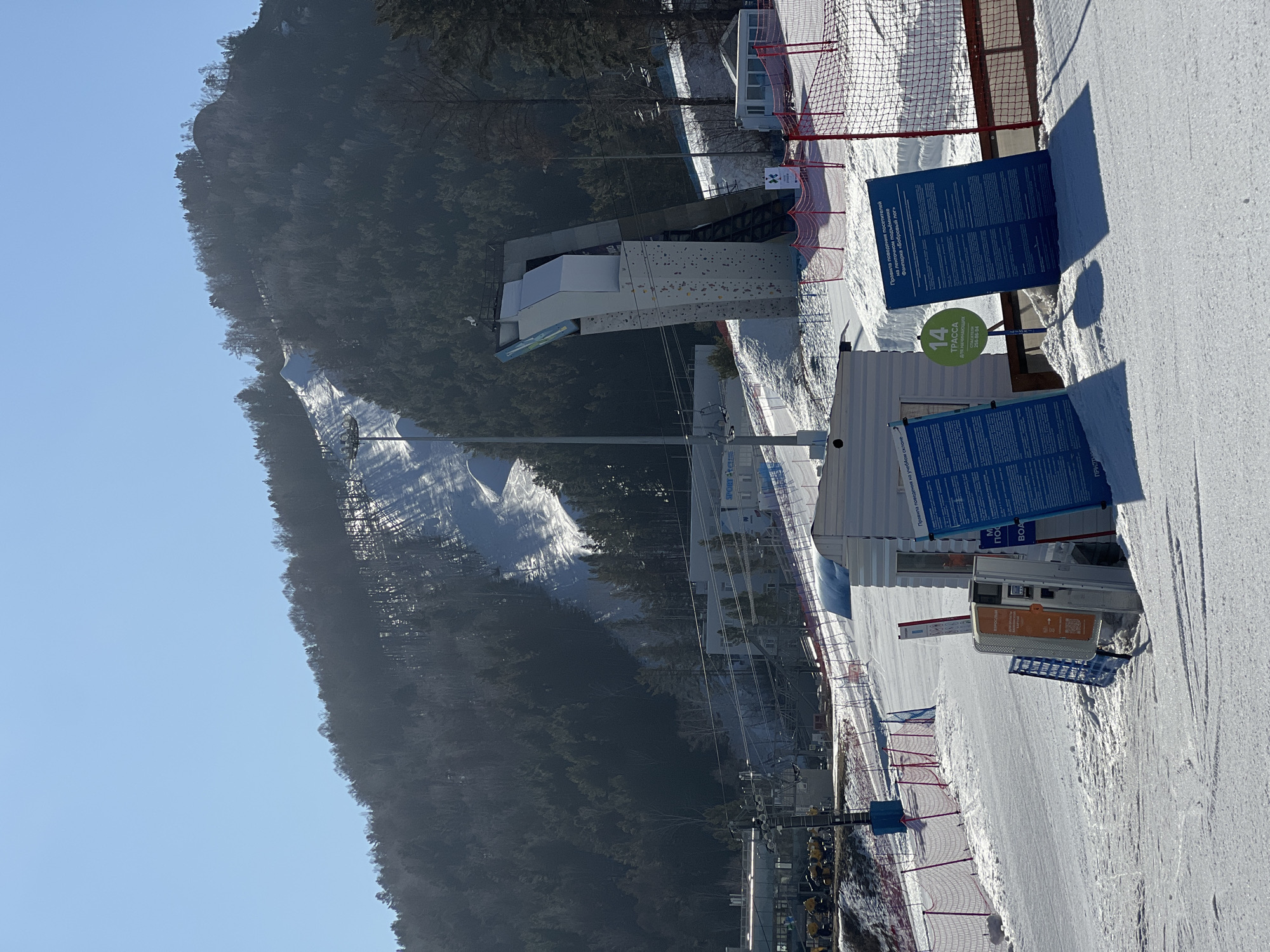 The image shows a winter scene at a ski resort, likely in a mountainous region. In the background, a snow-covered ski slope runs down a wooded mountain under a clear, blue sky. In the foreground, a flat, snow-covered area suggests the base of the slope.
Various structures are present, including a white building with what appears to be a climbing wall attached. Safety netting and fences are visible along the sides. A blue sign displays rules and information, including the number "14" in green, indicating a "TPACCA" (trail) for beginners. A vending machine or ticket kiosk is also noticeable. The scene is taken during daylight hours.