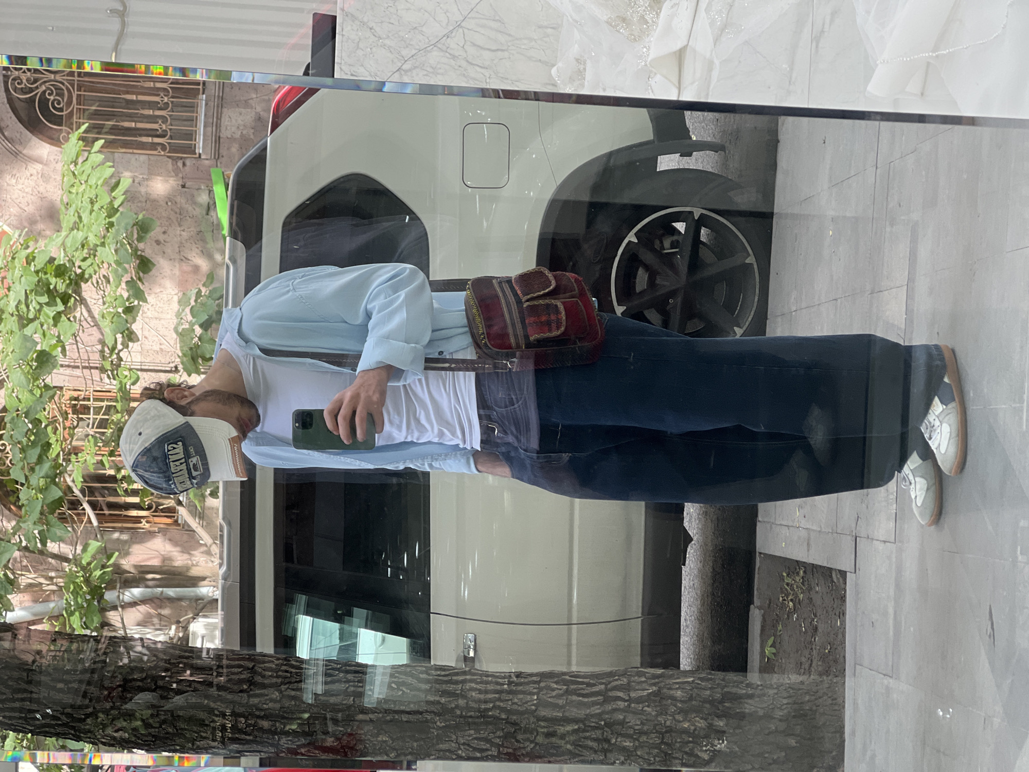 A man stands on a city sidewalk in Yerevan, Armenia, holding a smartphone. He is wearing a baseball cap, a light blue shirt over a white t-shirt, blue jeans, and sneakers. A small, patterned bag hangs across his body. He is standing in front of a window, which reflects a white vehicle with a visible tire and wheel. A tree is seen on the left side of the image, and a building with architectural details is visible above the tree. The sidewalk is made of gray paving stones.