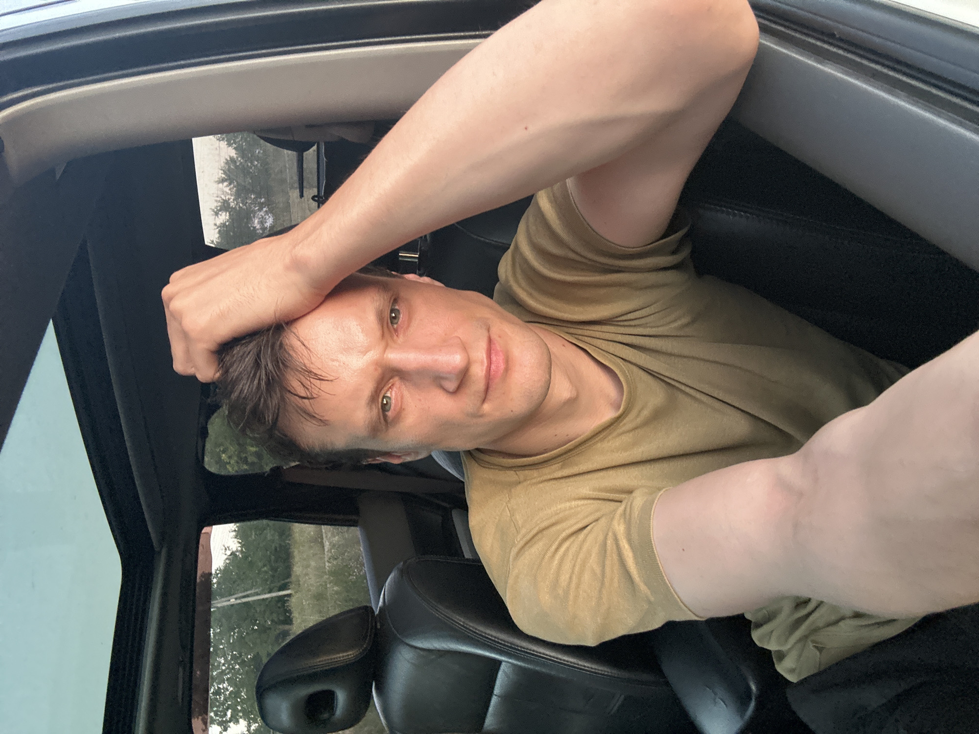 The image shows a man sitting in the driver's seat of a car, with the sunroof open. He is looking at the camera with his right arm resting on his head, and his left arm is resting on his lap. The man is wearing a light brown t-shirt. The interior of the car is visible, including black leather seats and a portion of the dashboard. Outside the car, trees and vegetation are visible, suggesting a rural or suburban setting. The lighting suggests it is late afternoon or early evening. There is no information in the image to suggest it is located in the city of Makiv, Ukraine.