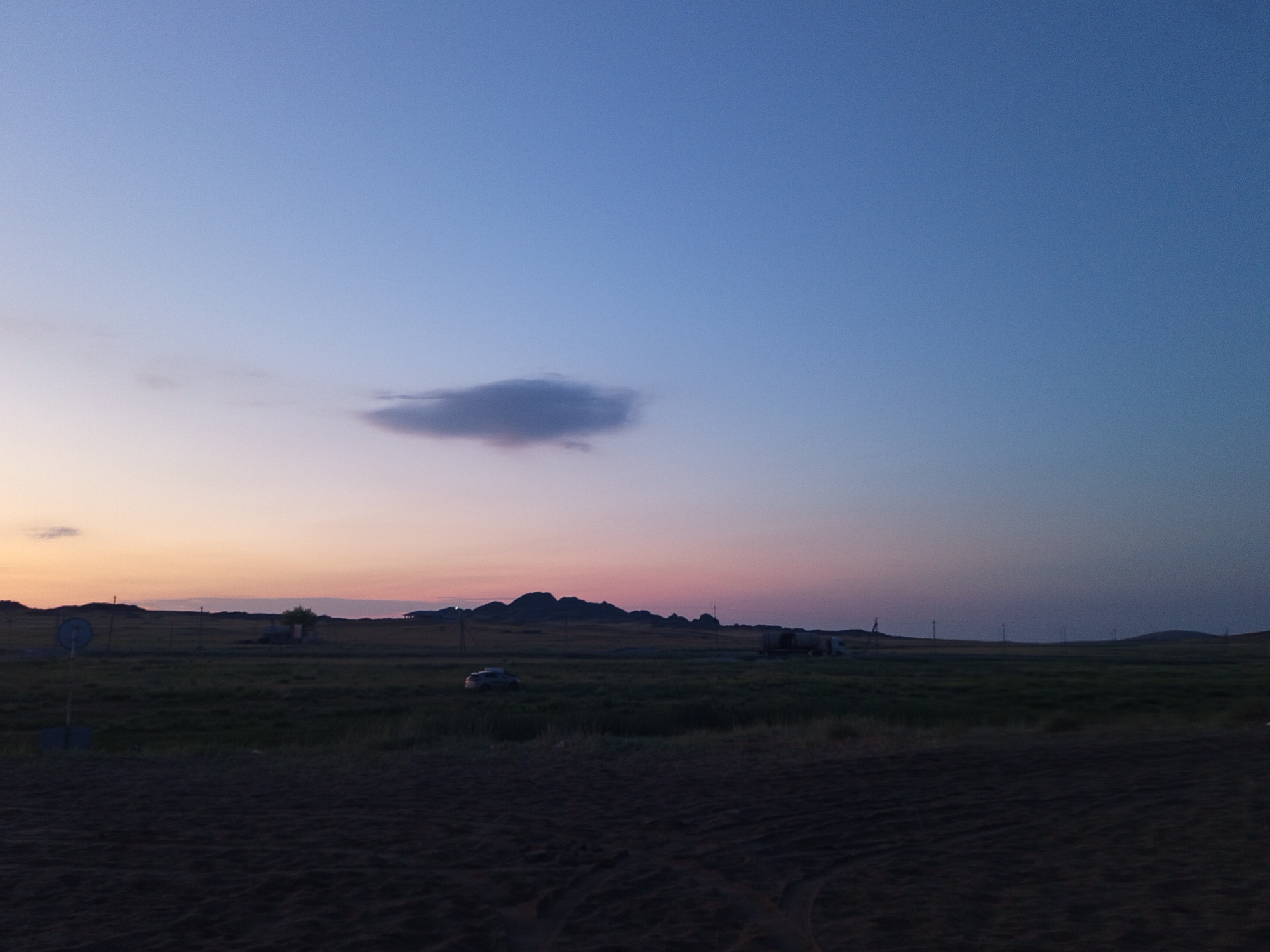 The image depicts a dusky, rural landscape, likely in the Bayanaul rural district of Kazakhstan. The sky transitions from a pale orange and pink near the horizon to a deep blue overhead, with a single, thin cloud illuminated by the setting sun. The terrain is predominantly flat, covered in dark green grass. In the mid-ground, a car is visible on the left, and further to the right, what appears to be a truck or a large vehicle is present. Several utility poles are scattered across the field, marking the presence of infrastructure. The foreground is a dark, rough, and textured surface, possibly soil or sand, showing tire tracks. Hills or low mountains are visible in the distance on the horizon, silhouetted against the fading light.
