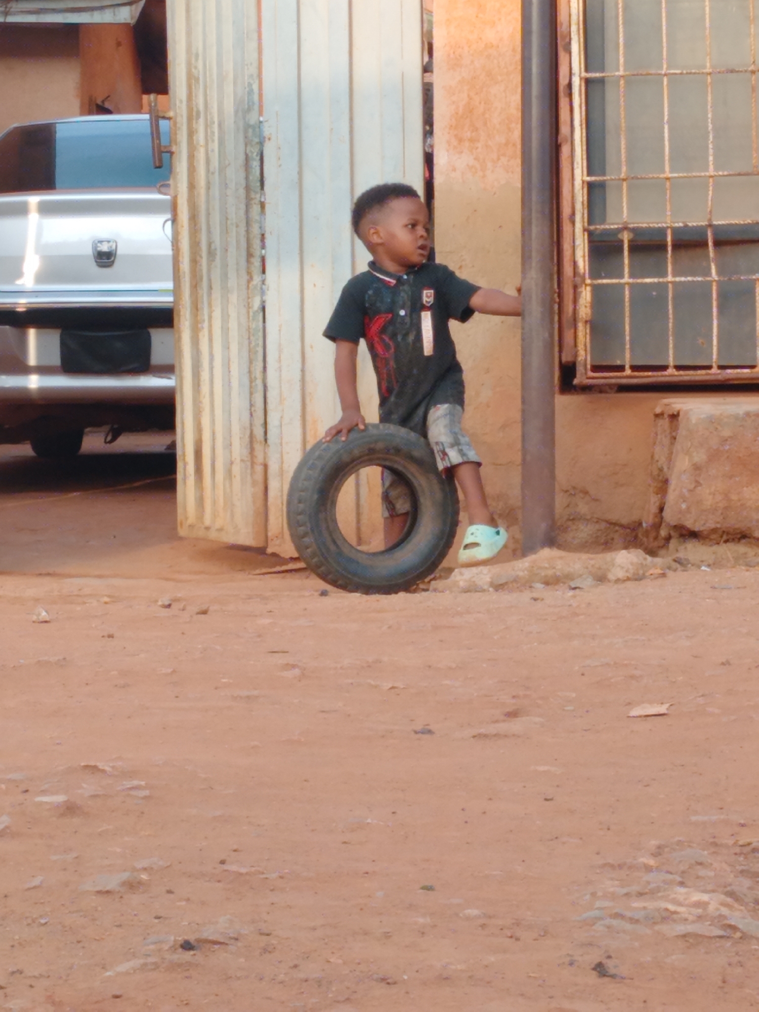 A young boy is playing with a tire in an outdoor, urban environment. He is leaning against the tire with his left hand. He is wearing a dark-colored short-sleeved shirt with a red graphic, plaid shorts, and light-colored sandals on his feet. The location appears to be near a building with a white corrugated metal door or wall and a window with metal bars. A silver car is partially visible behind the door. The ground is dirt and the environment suggests a low-income neighborhood, with signs of wear and tear on the building and surrounding area. The image was captured in Yelwa, Nigeria.