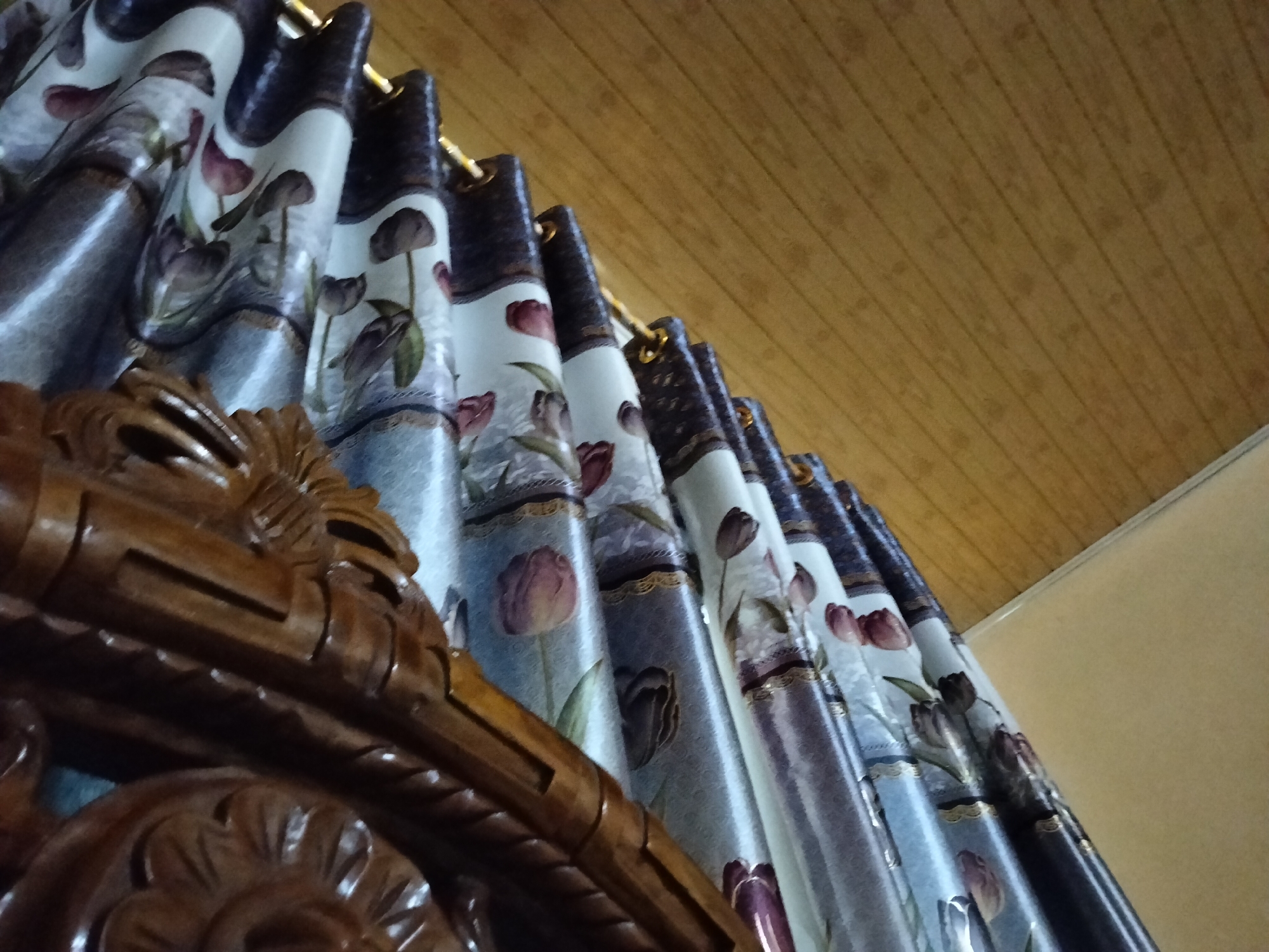 In Payagaboh, Indonesia, the image depicts a close-up, low-angle shot of a floral patterned curtain with a wood-grained ceiling overhead. The curtain, adorned with stylized tulips in shades of purple and pink against a light blue and white background, hangs in pleats. A decorative wooden element, possibly part of a furniture piece or architectural detail, is visible in the foreground, its dark brown, carved surface catching the light. The ceiling is constructed of light brown wooden planks arranged diagonally. The lighting suggests an indoor setting, possibly a room.