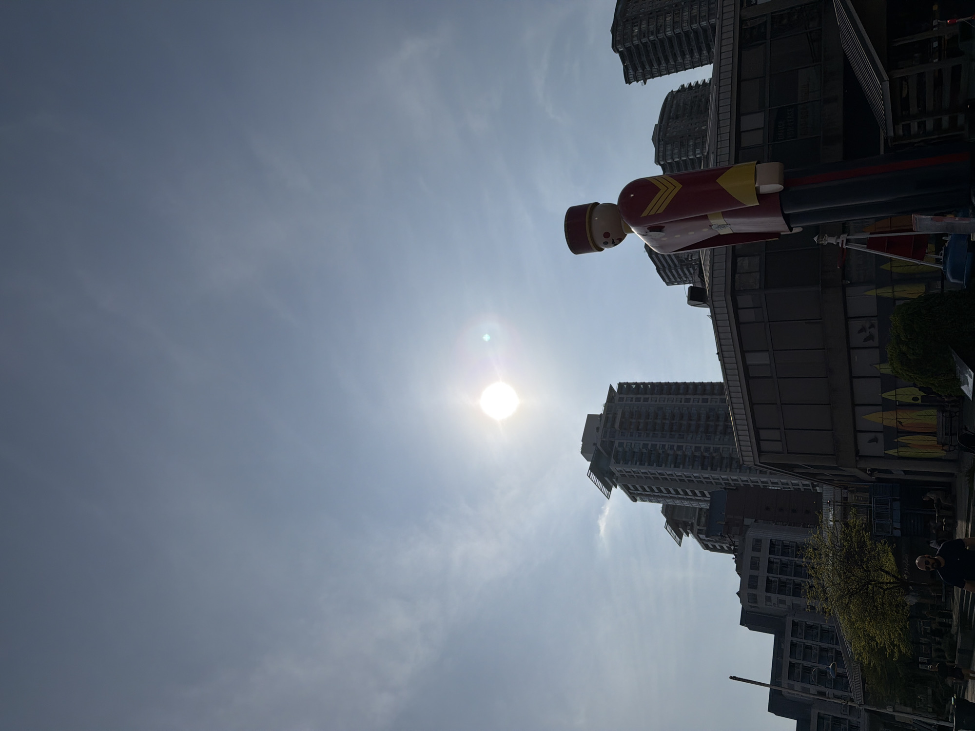 The image shows a cityscape with a large toy soldier statue in the foreground, viewed from a low angle, pointing towards the sky. The sun is visible in the sky, partially obscured by clouds. The buildings appear to be modern high-rise residential towers.
The location is New Westminster, British Columbia, Canada, based on the presence of the toy soldier statue, which is a local landmark.