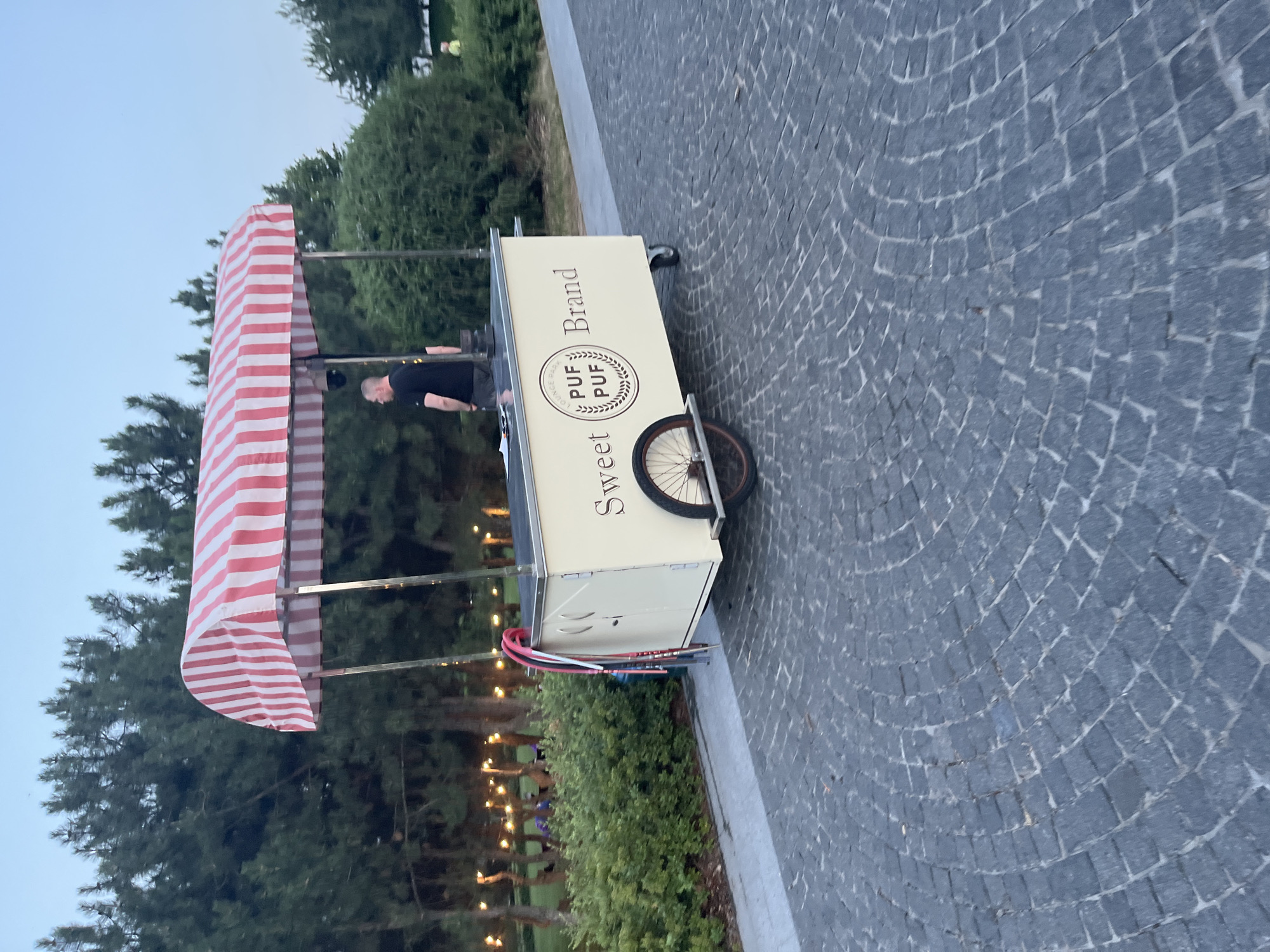 This is a medium shot, captured outdoors in the late afternoon or early evening, likely in the city of Dnipro, Ukraine. The foreground is dominated by a circular, cobblestone walkway. To the right, a green, manicured hedge runs parallel to the walkway. In the mid-ground, a cream-colored food cart with a red and white striped awning is parked. The cart has the words "Sweet PUF PUF Brand" inscribed on its side, and a round logo featuring a wreath and the text "LOUNGE BAR" is visible. A single spoked wheel is attached to the cart, and a person in a black t-shirt and dark pants is standing behind the counter of the cart, facing away from the camera. A security camera is mounted on one of the awning poles. In the background, a line of coniferous trees forms a dense forest, with string lights visible among them, suggesting a park or outdoor entertainment area. A person wearing a yellow shirt is visible in the far right background, near the hedge. The sky above is a pale blue, indicating twilight.