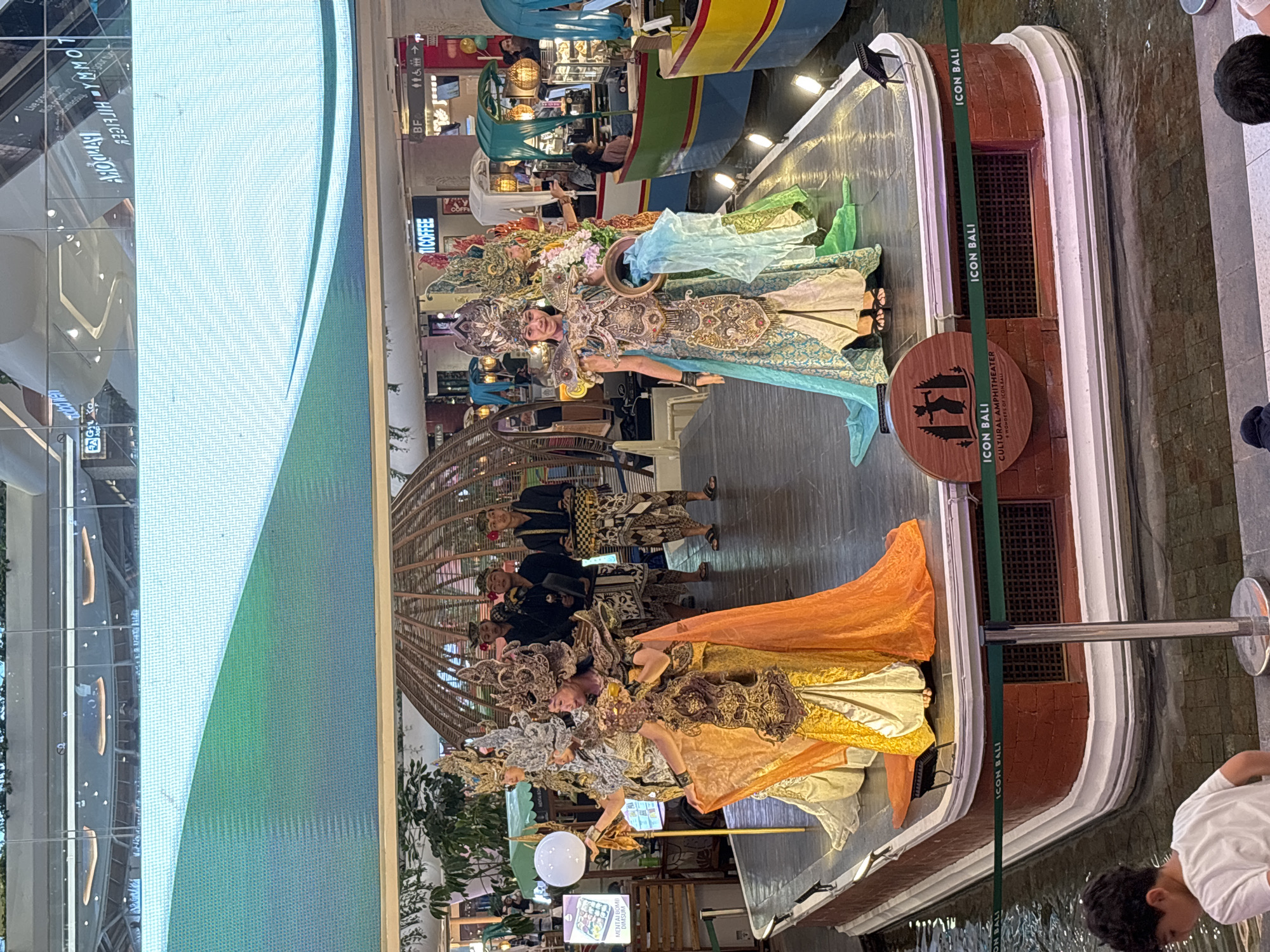 The image depicts a cultural performance at ICON BALI, a cultural amphitheater in Denpasar, Indonesia. The stage, which appears to be on a platform over water, features several performers in traditional Balinese attire. Two women in elaborate costumes with golden and blue dresses, headdresses, and ornate armor are positioned prominently. They are holding props; one holds a white orb, and the other holds a ring and flowing fabric. Behind them, four men in darker attire, also wearing traditional headwear, stand in a line. The background shows the interior of a shopping mall, with shops, lighting, and large digital screens visible overhead. Boats, possibly decorative elements or part of a water feature, are lined up to the right of the stage. A young boy in a white t-shirt is visible in the lower left foreground, looking down. The overall scene is a performance showcasing Balinese culture within a modern commercial setting.