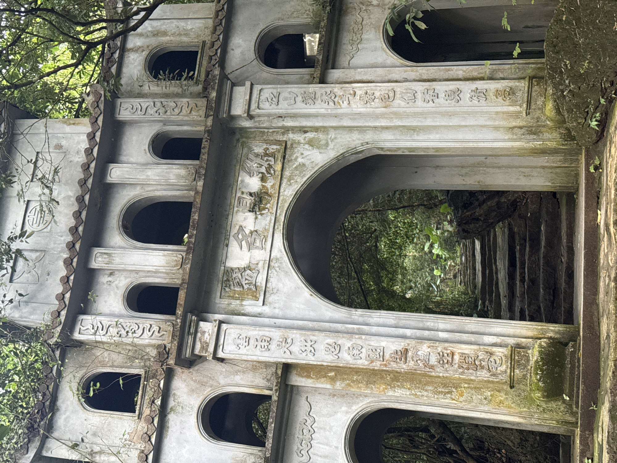 The image depicts a weathered, multi-tiered concrete structure with arched openings, likely an ancient gateway or temple entrance, situated amidst dense foliage. The structure is adorned with carved Chinese characters and decorative motifs. Moss and vines creep over the stone, indicating its age and integration with the natural environment. In the background, a stone staircase ascends into a verdant, overgrown forest. The scene suggests a historical or spiritual site, possibly within a rural or natural landscape, consistent with locations in Tây Hoa Lư Ward, Vietnam, known for its historical and cultural heritage sites. There are no people visible in the image.