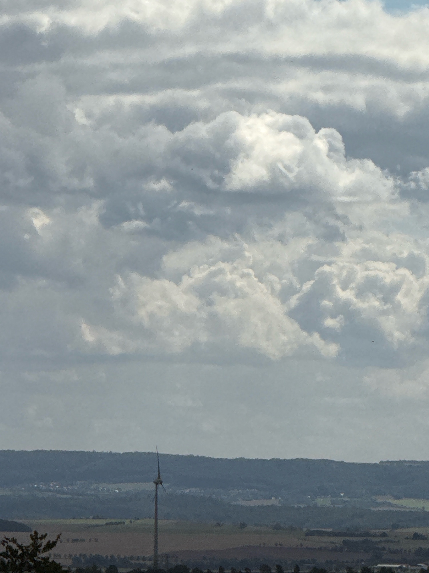 The image captures a distant view of a single wind turbine in a rural landscape, likely in the vicinity of Bad Sachsa, Germany. The turbine stands tall and slender against a backdrop of rolling hills and a hazy sky filled with dramatic, dense clouds. The lower portion of the image reveals fields and a sparse scattering of buildings nestled among the hills, suggesting a rural or semi-rural setting. The overall impression is one of a vast, open landscape under a cloudy, possibly overcast, sky.