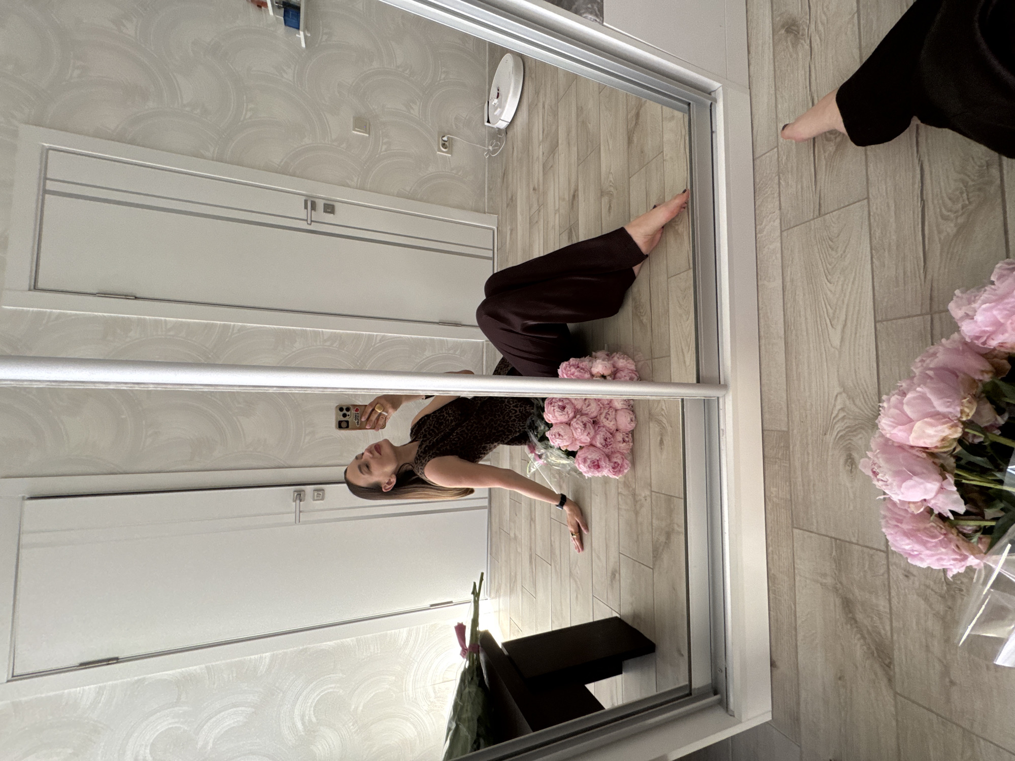 A woman is sitting on the floor in front of a mirrored wardrobe, taking a selfie. She has long brown hair and is wearing a sleeveless leopard-print top and dark brown wide-leg pants. She is holding a phone in her right hand and has a bouquet of pink peonies in front of her, with another bouquet of pink peonies and a long stemmed rose on a dark wood bench to her left. The room has light wood floors and white doors on the walls. A robot vacuum cleaner is visible on the floor in the background. The scene is indoors, likely in a residential setting.