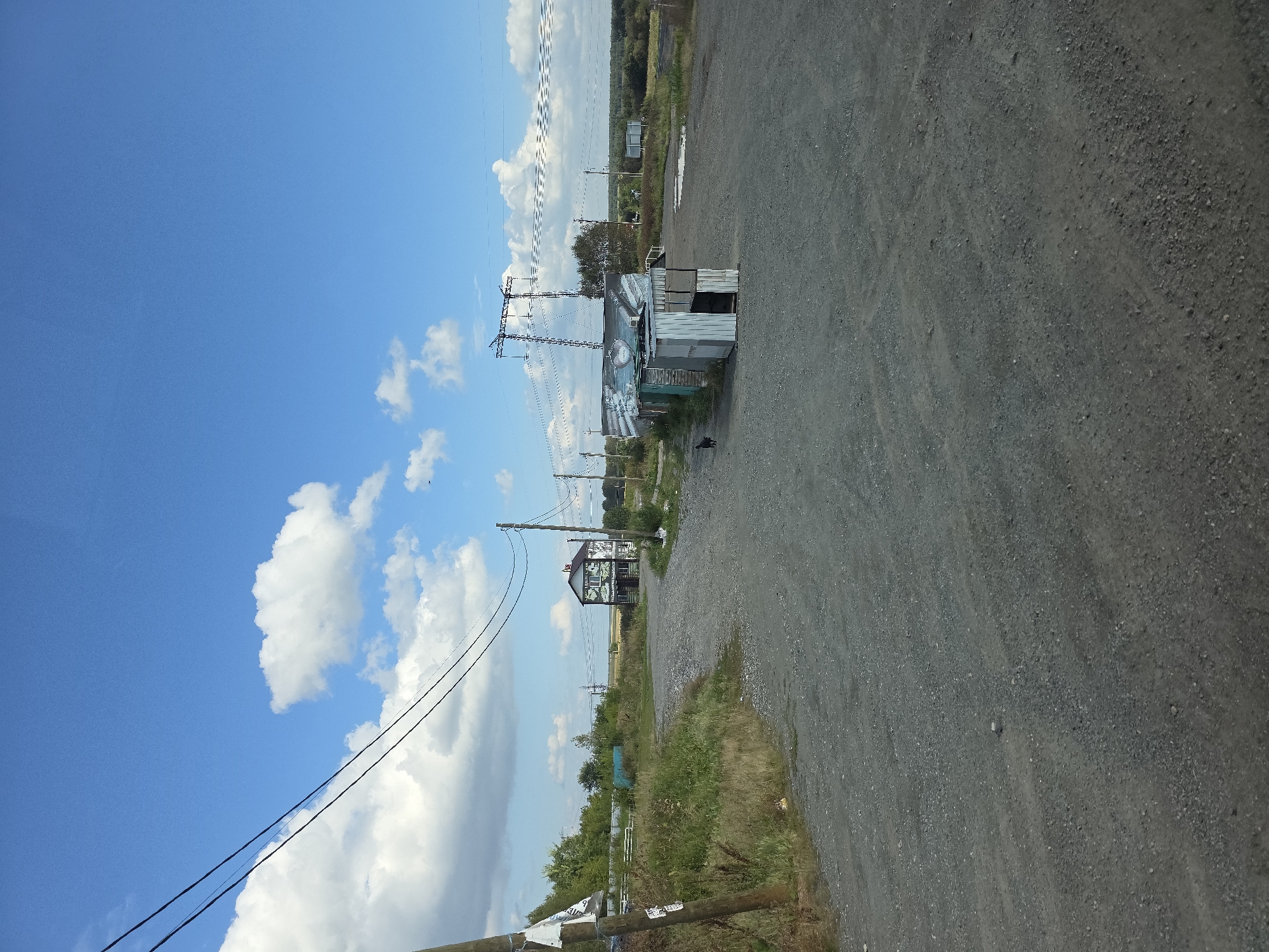 A wide, unpaved gravel road curves through a rural landscape under a bright blue sky dotted with fluffy white clouds. To the right of the road, a row of small, utilitarian structures, likely temporary sheds or market stalls, are clustered together. One of these structures is partially open, revealing an interior. A dark-colored dog stands near the front of these structures. Further down the road, a two-story building with a colorful mural on its side is visible. Overhead, power lines stretch across the frame, supported by wooden poles and a tall metal pylon. The surrounding terrain is a mix of overgrown grass and some scattered greenery. In the distance, a treeline and agricultural fields can be seen. The scene appears to be an outdoor market or a rural service area.