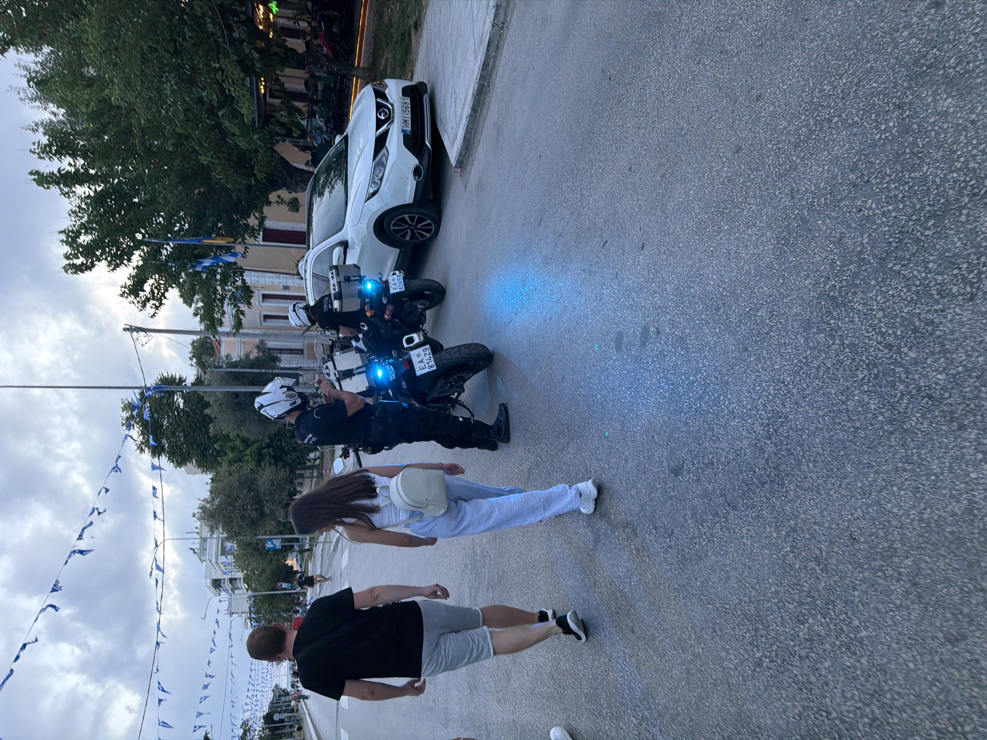 The image depicts a street scene in Alexandroupolis, Greece, under an overcast sky, likely in the late afternoon or early evening.
In the mid-ground, a police officer in uniform and a white helmet is positioned next to a black police motorcycle, which has its blue lights illuminated. A second police motorcycle is partially visible behind the first, also displaying blue lights. Both motorcycles have visible Hellenic Police (ΕΛ.ΑΣ.) license plates: "81959" is partially visible on one, and "81429" on the other. A white Nissan Qashqai car with the Greek license plate "HMM 5681" is parked nearby.
Two civilians are walking in the foreground: a woman with long brown hair wearing a white top, light blue trousers, and a white backpack, and a man in a black t-shirt and grey shorts. The street is decorated with numerous blue and white pennant flags, characteristic of Greek national colors, strung across the roadway. Trees and buildings line the street in the background, suggesting a bustling town or city environment. The scene appears calm and shows everyday street activity.
