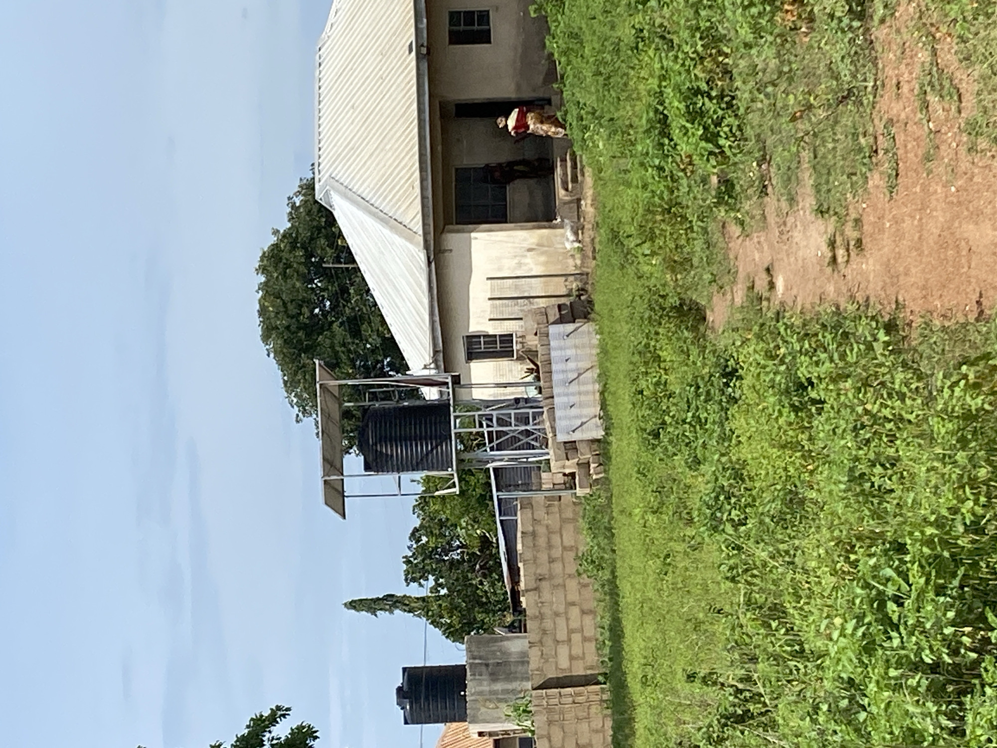 In the city of Unknown, Unknown, a wide shot captures a sunlit outdoor scene with a dirt path leading through lush green vegetation towards a modest, light-colored house. Two large black water tanks are visible; one stands on a concrete block structure to the left, and another is elevated on a metal frame with a solar panel on top, positioned to the right of the first tank. A brick wall runs horizontally across the mid-ground. Two figures, one appearing to be a child and another an adult wearing a patterned garment, stand near the entrance of the house, which features a covered porch and windows. Trees with green foliage are present in the background, and the sky is a pale blue with scattered clouds.
