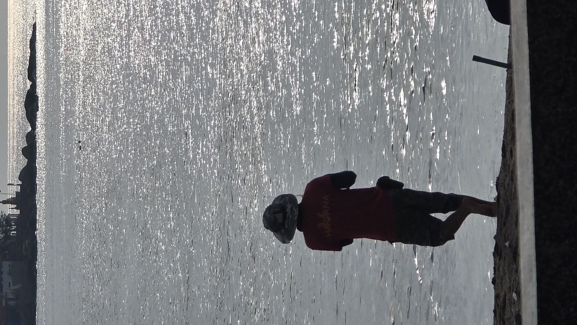 A person is standing on the edge of a concrete embankment overlooking a body of water. The person is wearing a red shirt with the word "HABANA" printed on the back, shorts, and a hat. The water appears to be shimmering due to sunlight. In the distance, along the left side of the image, there is land with some buildings and other structures visible. The location is Nha Trang Ward, Vietnam.