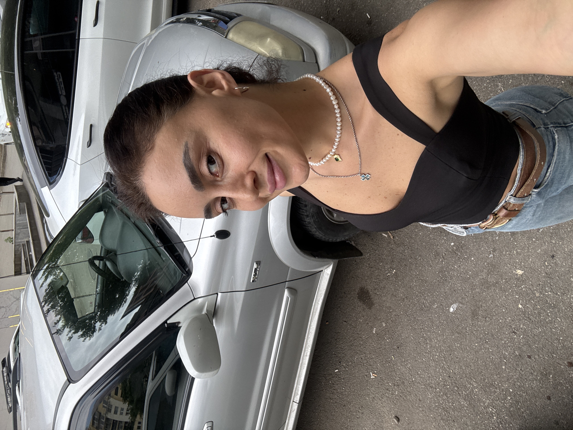 A young woman, with dark hair pulled back and wearing a black top and jeans, takes a selfie outdoors. She is standing next to a silver SUV. In the background, a white Tesla can be seen. The scene appears to be on a street in Tbilisi, Georgia, judging by the architecture and the presence of a typical Georgian street. The ground is asphalt, with some scattered debris. The woman is wearing a pearl necklace and a pendant.