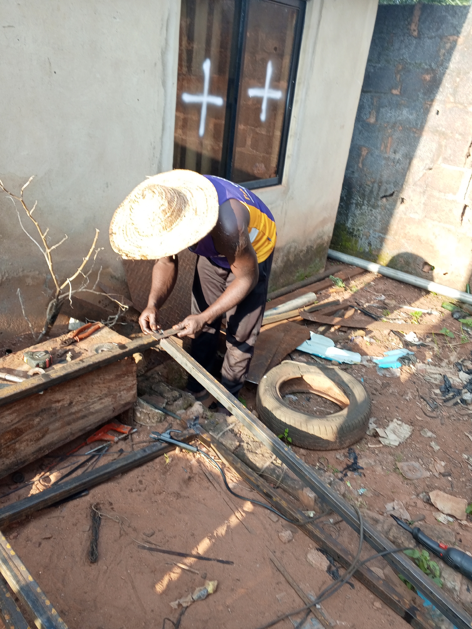 The image depicts an outdoor scene in Ankpa, Nigeria, where a person wearing a straw hat and a purple and yellow shirt is working on metal framing. The person is bent over, holding a piece of metal and what appears to be a measuring tape or ruler, engaging in what looks like fabrication or assembly. Various tools, including pliers and a saw, are visible on a wooden surface nearby. The ground is a mixture of red earth and debris, with scattered metal pieces, a tire, and some vegetation. A building with a window marked with two white crosses is visible in the background, adjacent to a brick wall. The lighting suggests it is daytime, with shadows cast by the building.