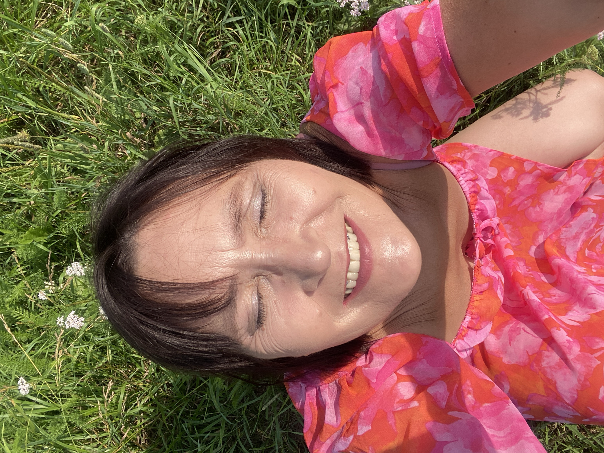 A woman with dark hair and closed eyes lies on her back in a grassy field. She is wearing a pink and orange floral dress. The sun shines on her face, highlighting her smile. The field appears to be in a rural or park setting, with green grass and some small white flowers. There is no visual information to place this image in Novokuznetsk, Russia.