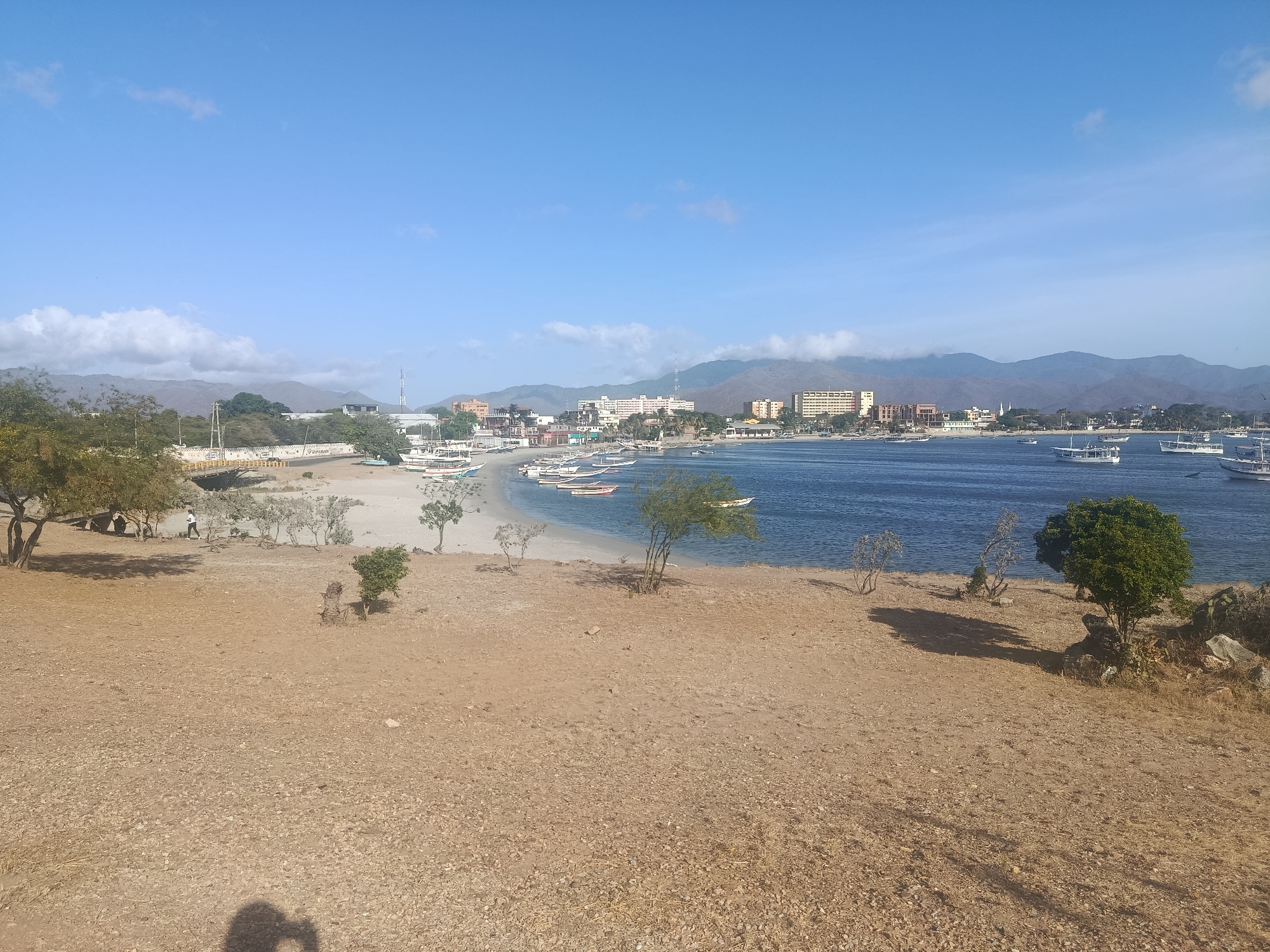 The image depicts a coastal scene in Juan Griego, Venezuela. The foreground features dry, barren land with scattered sparse vegetation. A sandy beach lies between the dry land and the water. Several small boats are anchored in the calm, blue water. In the background, a coastal town is visible, with buildings of varying heights and styles. Mountains rise in the distance, partially obscured by clouds. The sky is a clear blue with scattered clouds. A bridge is seen in the left background, next to a building. There are a few people seen on the beach by the bridge. The shadow of the photographer is cast in the lower center of the scene.