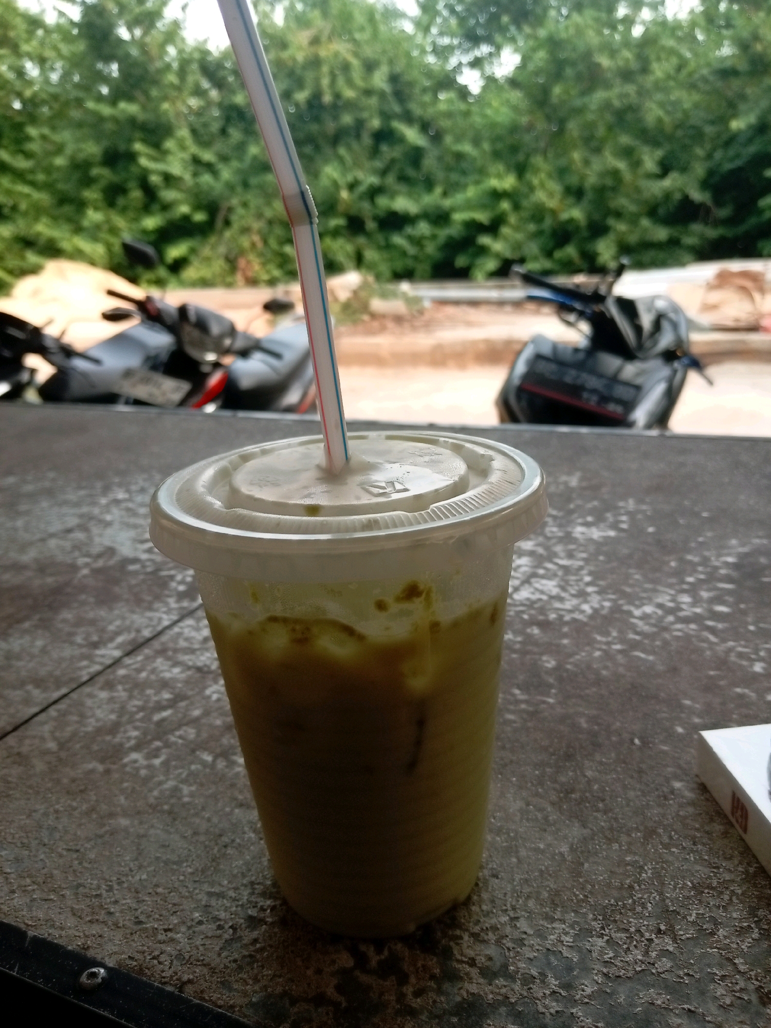 A plastic cup containing a green beverage with ice and a straw rests on a speckled dark gray surface, likely a table. The cup is partially filled, with some condensation visible on its exterior. In the background, out of focus, are two parked motorcycles and a dense wall of green trees. The scene appears to be outdoors, possibly at a roadside stall or cafe in Batam, Indonesia.