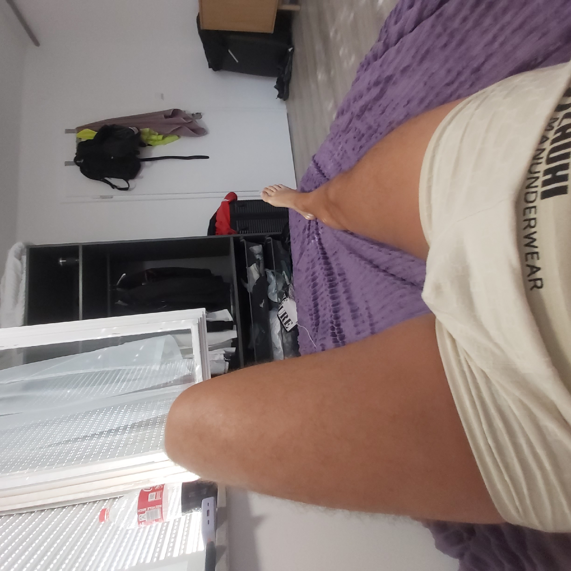 The image depicts a first-person perspective of a person lying on a bed with a purple textured blanket. The person's legs, clad in light-colored underwear with visible text "MAHNUDERWEAR," are prominently featured. The underwear has a ribbed texture. To the left, a windowsill is visible with blinds and a partially obscured bottle of soda. Further left, a white curtain hangs. In the background, a dark grey wardrobe with shelves containing various items, including clothing, is present. To the right, a white wall displays a backpack and towels hanging on a hook. A dark piece of luggage and a wooden cabinet are also visible. The floor appears to be light-colored wood. The scene is indoors, likely a bedroom. There is no specific indication that the location is in the city of Gärtringen, Germany.