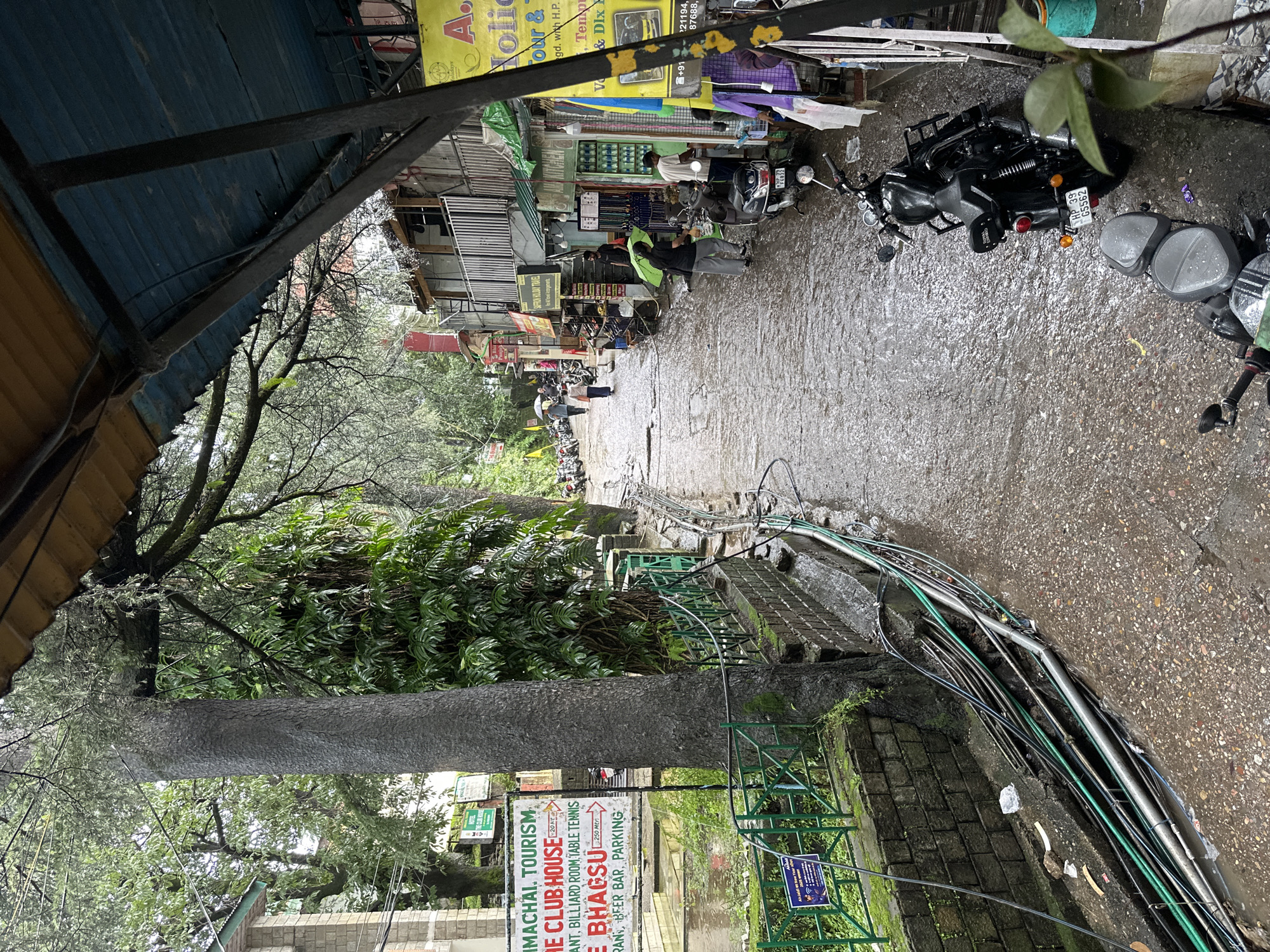 The image shows a narrow, wet street in what appears to be a market area with shops lining one side. The street is paved with a rough, textured material and is covered in a shallow layer of water, suggesting recent rain. Several motorcycles are parked on the side of the street, and a few people are walking, some with umbrellas. The buildings on the right are two or three stories high and have balconies. Shops display various goods, including clothing and what looks like souvenirs. A large tree with a thick trunk is prominent on the left side of the image, with green fencing and a stone wall running along its base. There are also many exposed electrical wires and pipes running along the ground and on the wall. Signage is visible, including one that mentions "Himachal Tourism" and "The Club House." The overall atmosphere is that of a bustling, somewhat unkempt, urban street scene during or immediately after rainfall.