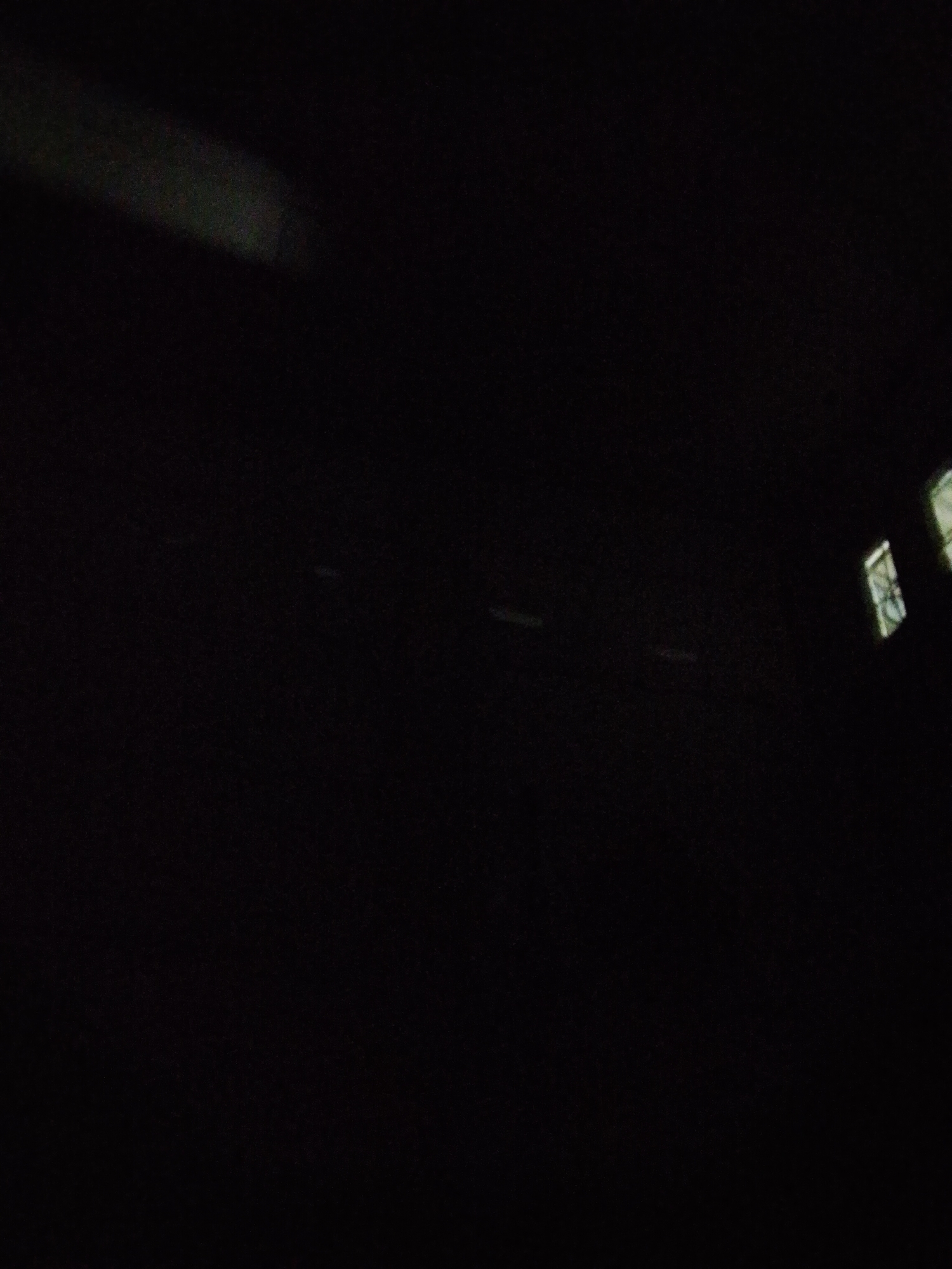 The image appears to be taken inside a dark tunnel or covered passageway in Bukit Ketok, Indonesia. There are faint, blurry lights at the far right, possibly indicating the tunnel's exit or openings to the outside. The rest of the space is shrouded in darkness, with only the faintest suggestion of shapes along the walls.