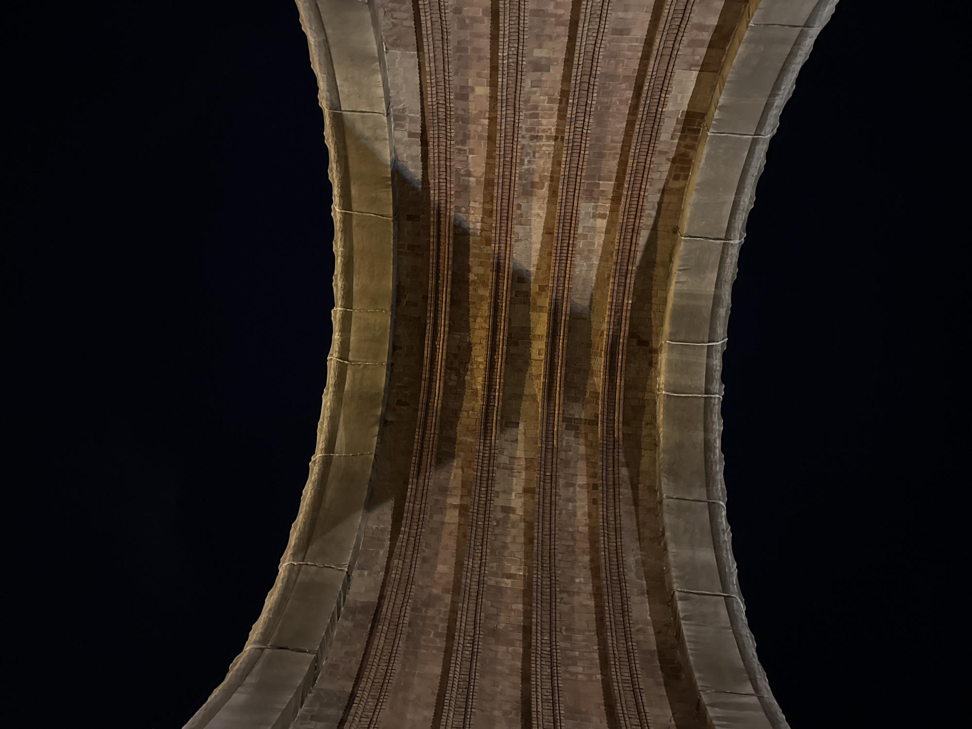 The image depicts the underside of the Pont de Vallcarca, a viaduct in Barcelona, Spain. The perspective is from below, looking upwards at the structure against a dark night sky. The viaduct is constructed of brick with stone or concrete supports along the edges. The roadbed is marked by parallel lines which likely represent railway tracks. The structure curves inwards towards the center of the frame, creating a narrowing effect. The lighting is uneven, casting shadows that accentuate the depth and texture of the brickwork.