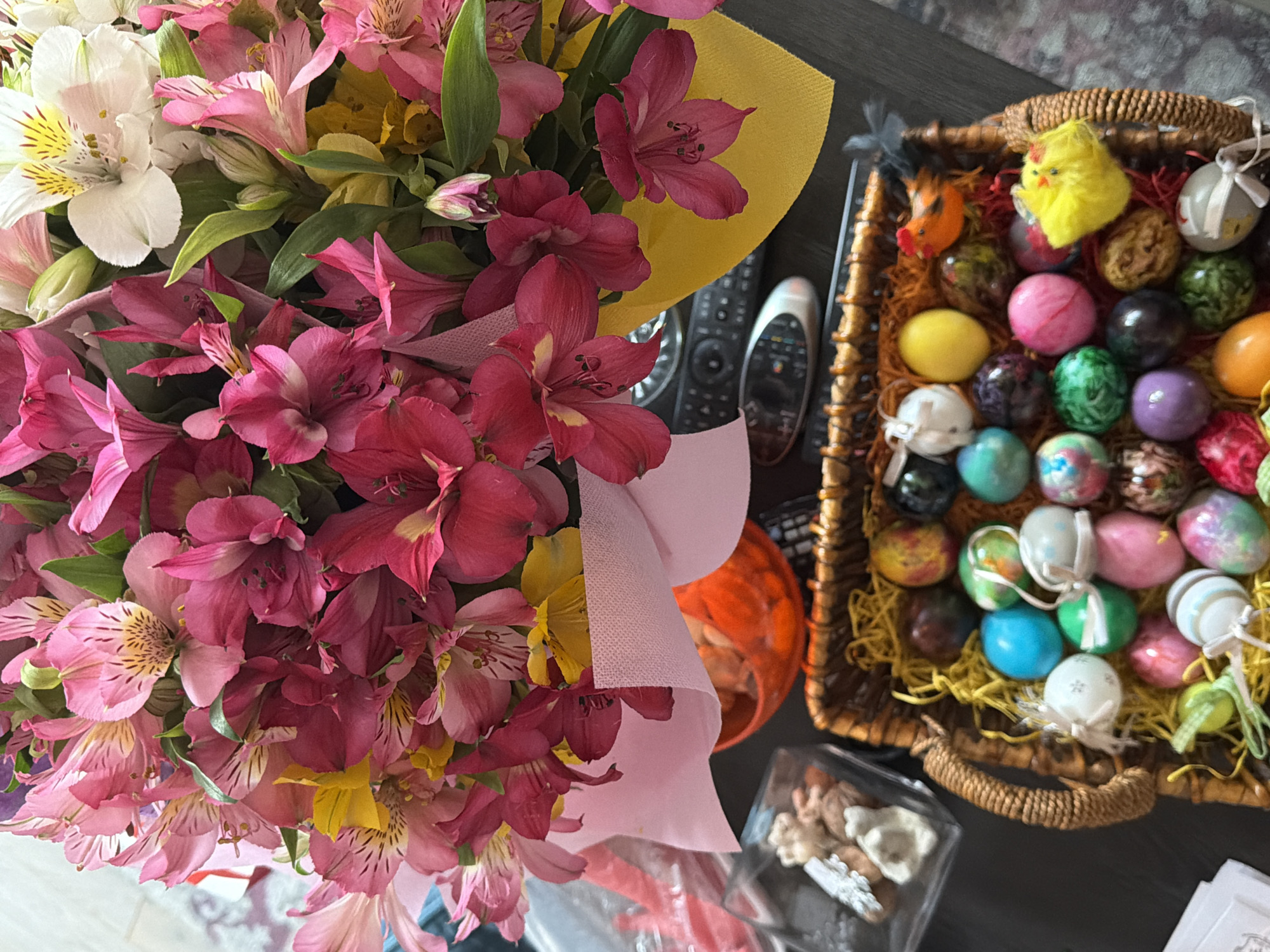 The image depicts a still life arrangement on a dark, possibly wooden surface. A large bouquet of pink, white, and yellow alstroemeria flowers dominates the left side. To the right, a wicker basket filled with decorated Easter eggs is visible, along with small yellow and orange decorative chicks. Two remote controls are placed near the basket. Additional items include a transparent container with what appears to be potpourri and an orange container of unknown contents. The environment suggests an interior setting, and no people are visible. There's no indication of Sofia, Bulgaria.