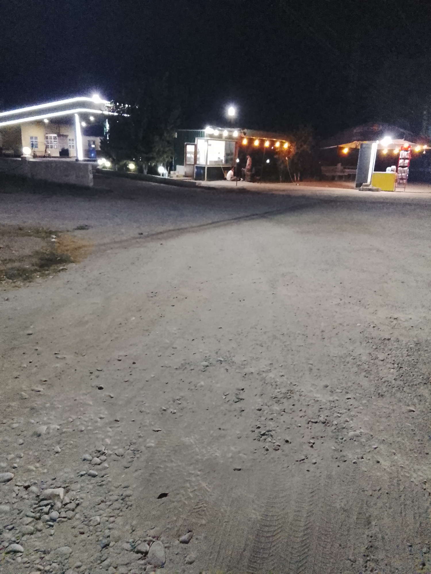 The image captures a nighttime scene in the city of Sabir Rakhimov, Uzbekistan, likely a roadside rest stop or a marketplace area. The foreground is dominated by a rough, unpaved ground surface with scattered stones and tire tracks, indicative of a dusty, rural or semi-rural environment.
In the background, several illuminated structures are visible. To the left, a building with a covered canopy, possibly a gas station or a small shop, is lit by strings of white lights along its edges. Windows are visible, and it appears to be open for business or operation. Further to the right, a smaller, open-air food stall or kiosk is illuminated by a string of warm, yellow lights. Two individuals are seated at this stall. Adjacent to this, another structure with a thatched or fabric roof is also lit, suggesting a commercial or recreational area. The overall lighting suggests a warm, inviting atmosphere despite the darkness of the night. The background trees are silhouetted against the dark sky.