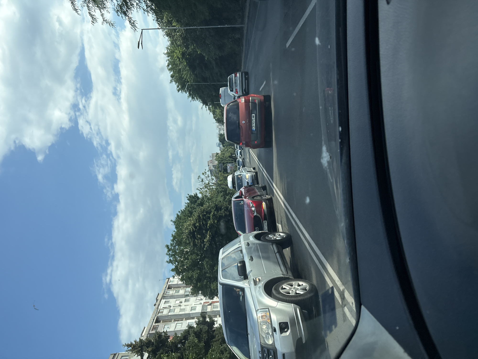 The image shows a traffic jam on a sunny day in Burgas, Bulgaria. In the foreground, a silver Land Rover Freelander is stopped in traffic. To its right, a red Renault Scenic is also stopped, followed by a white van and a white Audi. In the distance, more cars and buildings are visible, along with trees and a blue sky with white clouds. A bird can be seen flying in the sky. The scene appears to be on a multi-lane road.