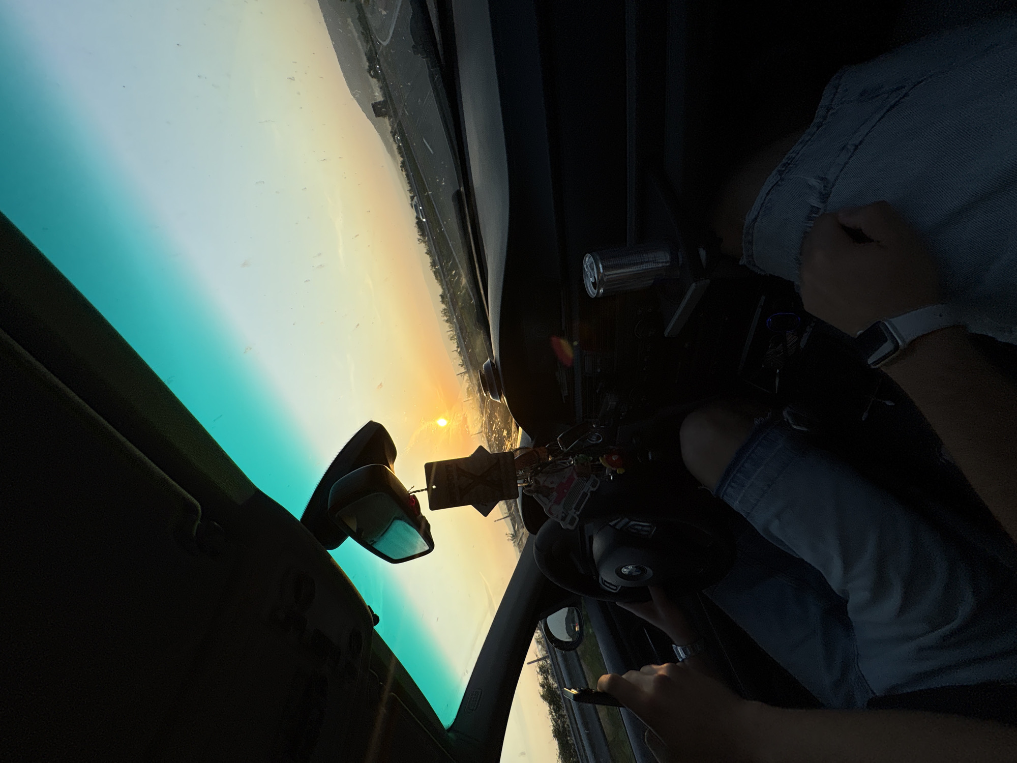 The image captures the interior perspective from a vehicle traveling on a multi-lane road in Devnya, Bulgaria, during either sunrise or sunset. The sun, a distinct orange orb, is low on the horizon through the front windshield, casting intense light into the vehicle's cabin. The upper section of the windshield exhibits a green-blue tint, and the exterior surface displays numerous small specks of dust or dirt.
Two occupants are visible within the vehicle. The individual in the driver's seat, on the left, has their left hand holding a black smartphone and their right hand partially gripping a black multi-function steering wheel with a visible emblem. This person is wearing blue denim shorts or jeans. A second individual, presumed to be in the front passenger seat, has their left arm visible on the right side of the frame, wearing a white smartwatch. Their lower torso, clad in blue denim shorts or jeans, is also visible.
The vehicle's interior features a dark dashboard. A silver cylindrical object, resembling a beverage can, is situated in a console or holder on the passenger side. A rearview mirror is centrally positioned, from which a black air freshener with a white 'X' design hangs, accompanied by a miniature cartoon car and several other small decorative charms. The interior roof liner and sun visors are dark in color.
The exterior scene depicts a highway or major road extending into the distance, bordered by guardrails and sparse vegetation. Distant hills or low mountains are visible on the right horizon, and other vehicles appear as distant forms on the roadway.