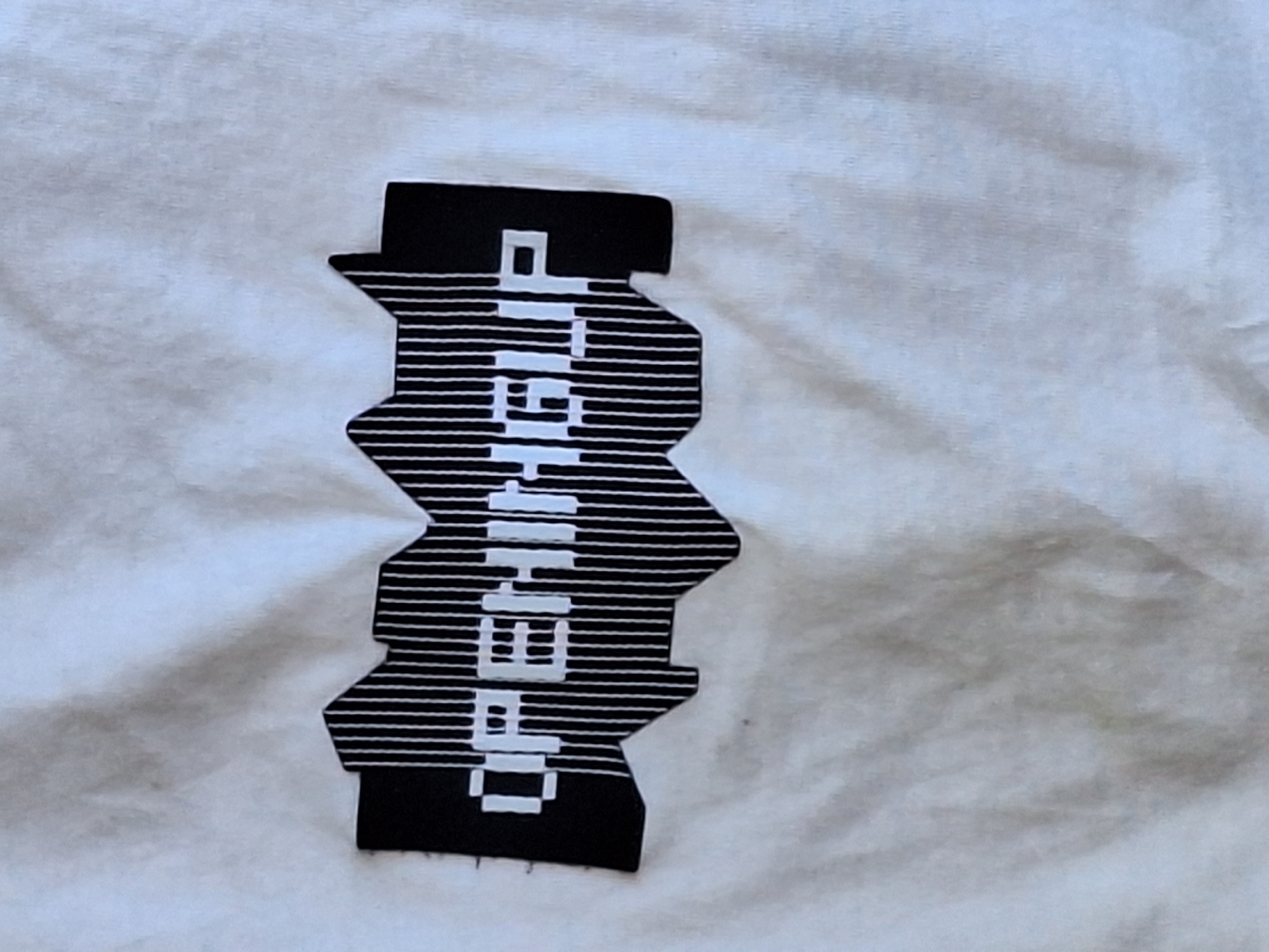 The image displays a close-up, overhead shot of a white t-shirt with a black and white graphic design. The graphic is a stylized, jagged shape with vertical black lines filling the interior. In the center of this graphic, the word "OPENINGUP" is printed in white, pixelated letters. The t-shirt exhibits wrinkles and folds, indicating it is likely being worn or has been stored. There is a faint, unidentifiable dark mark on the t-shirt below and to the left of the graphic. The background is entirely composed of the white fabric of the t-shirt. There are no people, other objects, or environmental features visible that would allow for the identification of a specific location in Varna, Bulgaria.