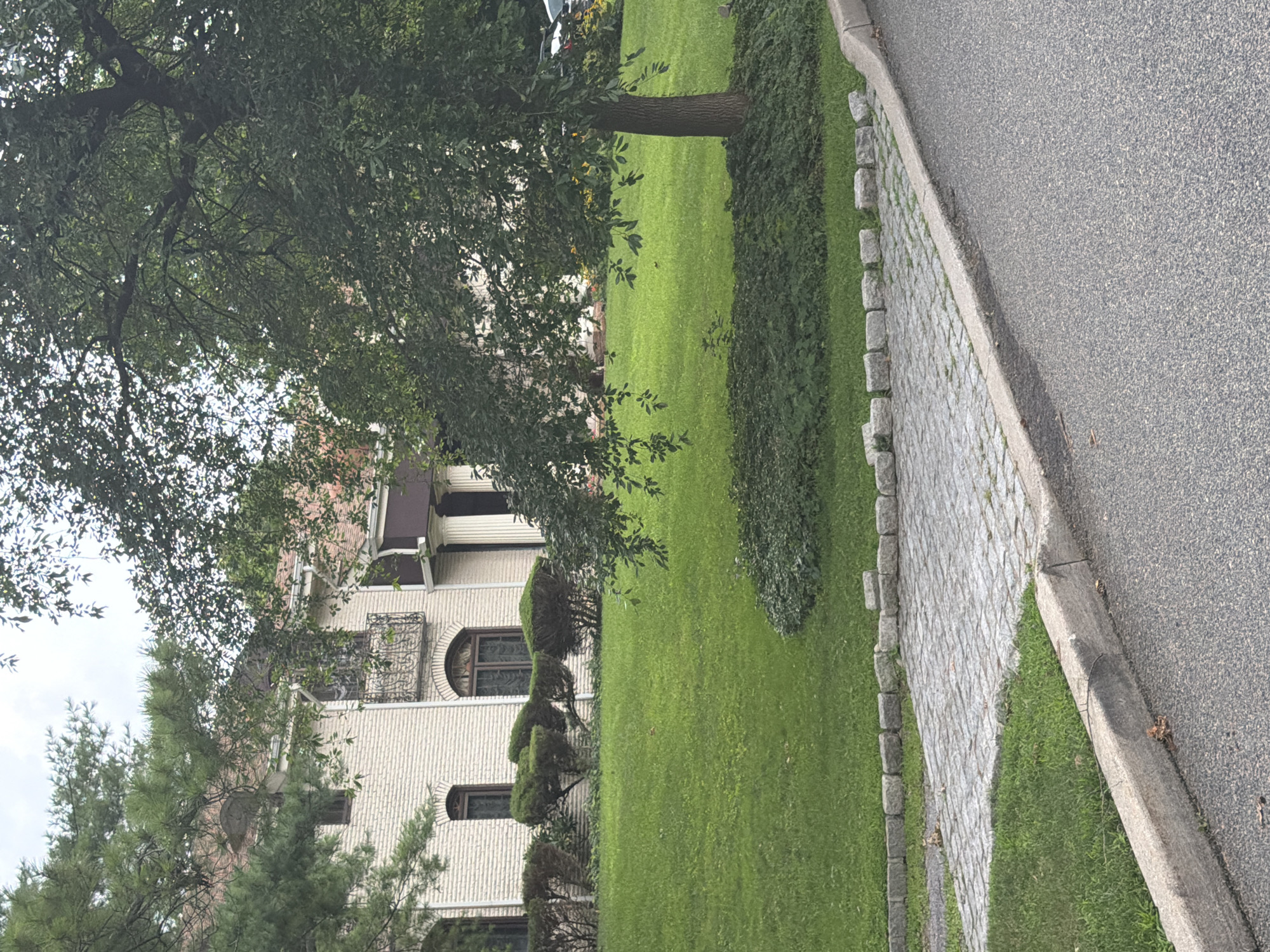 The image depicts a residential scene in the Village of Great Neck, USA, featuring a large, light-colored brick house with a red-tiled roof, partially obscured by mature trees and lush green landscaping. In the foreground, a paved driveway made of cobblestones transitions to a grey asphalt road, bordered by a concrete curb. The house exhibits architectural details such as arched windows and ornate balconies. The yard is well-maintained with a vibrant green lawn and neatly trimmed bushes. A large tree with dense foliage stands prominently on the right side of the frame, casting shadows on the lawn. The overall environment suggests a suburban setting with well-established properties. There are no people visible in the image.