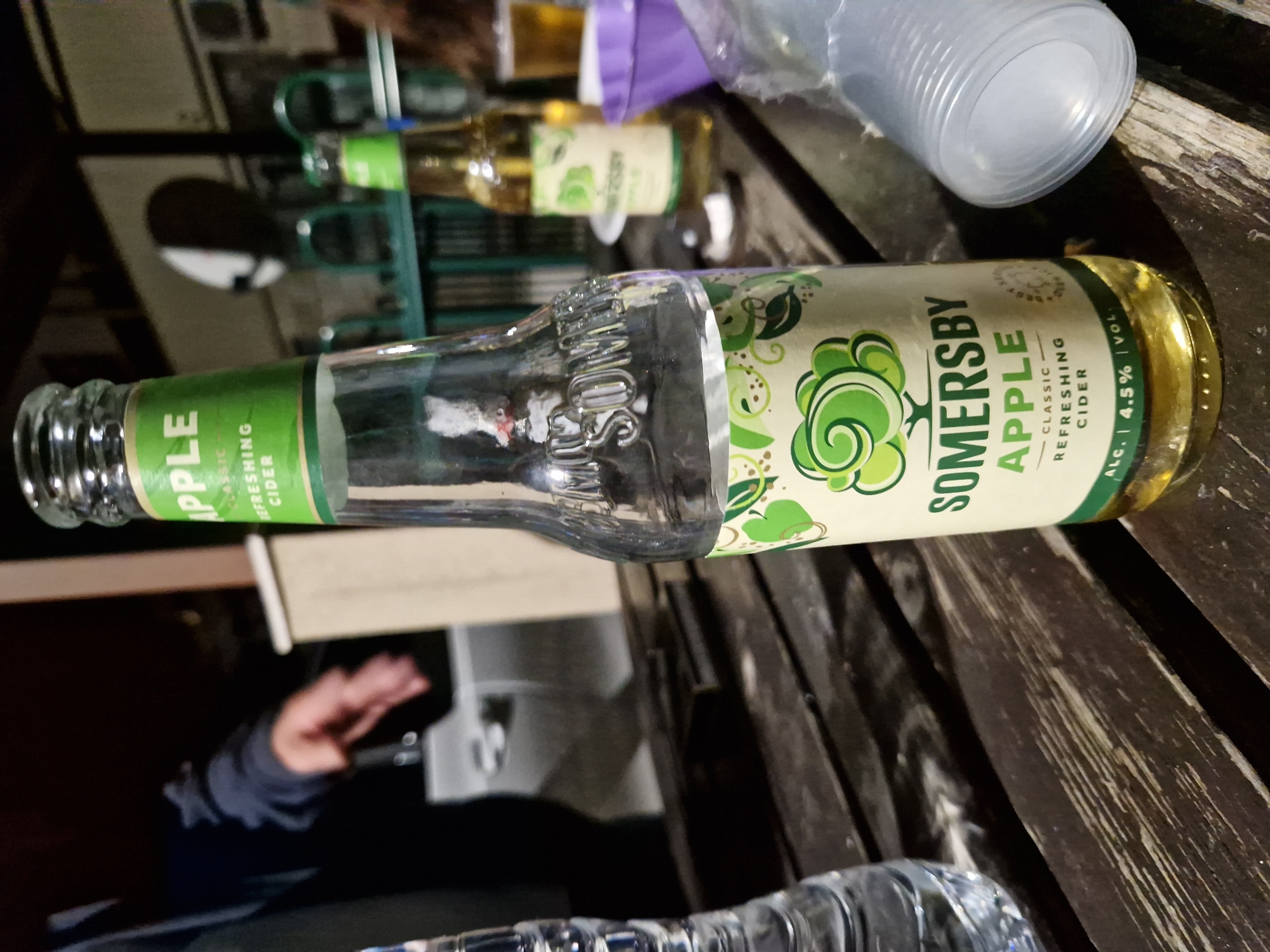 A clear glass bottle of Somersby Apple cider is positioned on a dark wooden picnic table. The bottle has a green label with white text, featuring the brand name "SOMERSBY" embossed on the glass. To the left of the cider bottle is a partially visible plastic water bottle. In the background, another bottle of Somersby cider, this one with a lighter colored liquid, is visible, along with a purple plastic cup and a clear plastic cup, suggesting an outdoor gathering. The scene appears to be an outdoor setting, possibly a beer garden or patio. The lighting suggests it is nighttime or late evening. No specific landmarks that would definitively place it in Sofia, Bulgaria are visible.