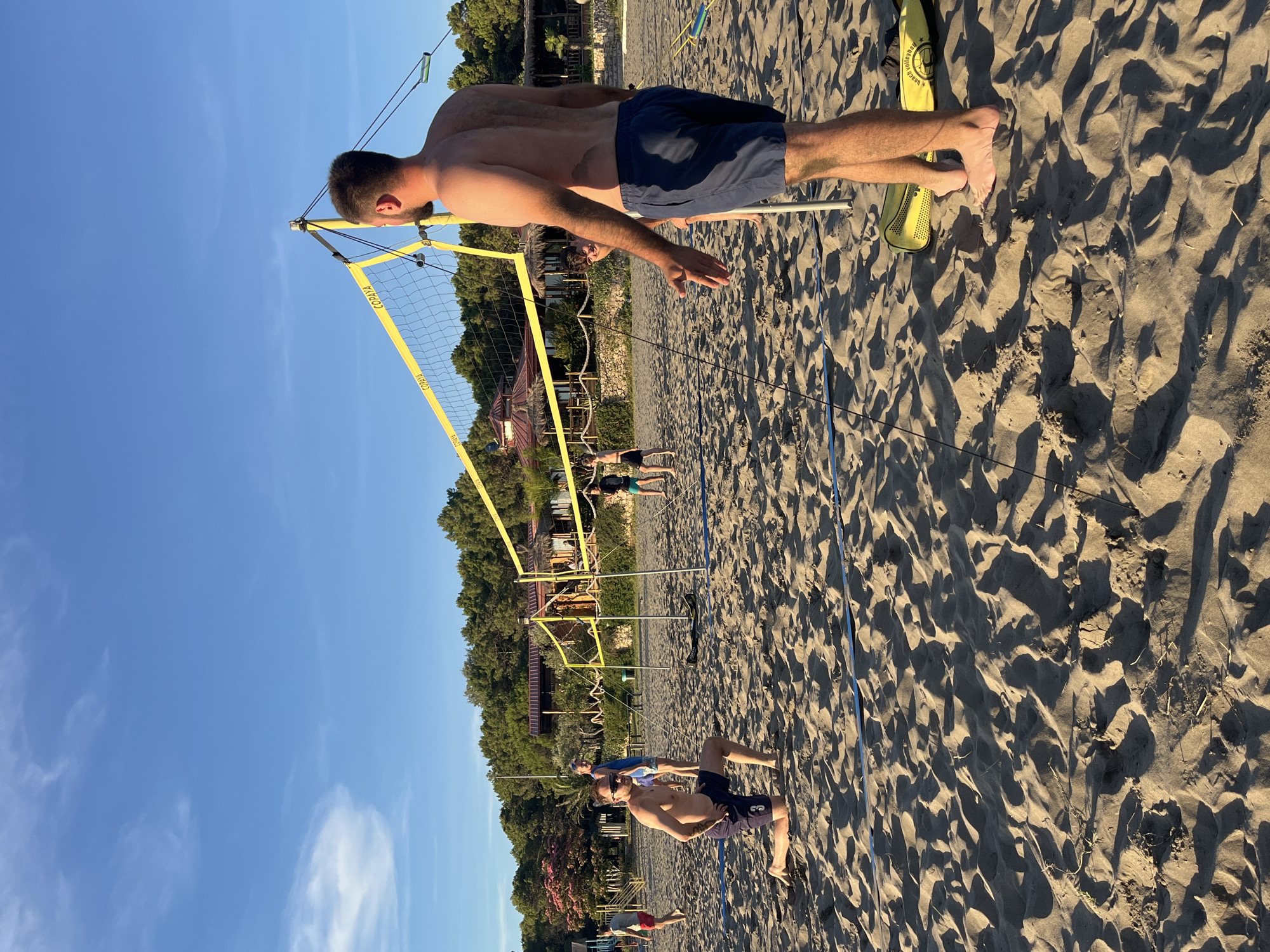 This is a medium-shot, eye-level, outdoor photograph taken on a sandy beach in Pistula, Montenegro. The scene depicts a group of people playing beach volleyball on a sunny day. The sand is light brown and textured with footprints and shadows. A bright yellow volleyball net is set up across the scene, with yellow poles supporting it. In the foreground, to the right, a shirtless man wearing navy blue shorts stands with his back to the camera, facing the net. His right arm is slightly extended, and his left hand is held up, possibly in a gesture of communication or instruction. To the left, in the mid-ground, a shirtless man in navy blue shorts and sunglasses is kneeling on the sand, looking towards the net. He has the number '3' visible on his shorts. In the background, other people are visible playing volleyball. Beyond the volleyball court, there are trees and some buildings with red roofs, suggesting a coastal resort area. The sky is a clear, bright blue with a few wispy white clouds. The overall impression is one of a casual, recreational activity on a warm, sunny day.