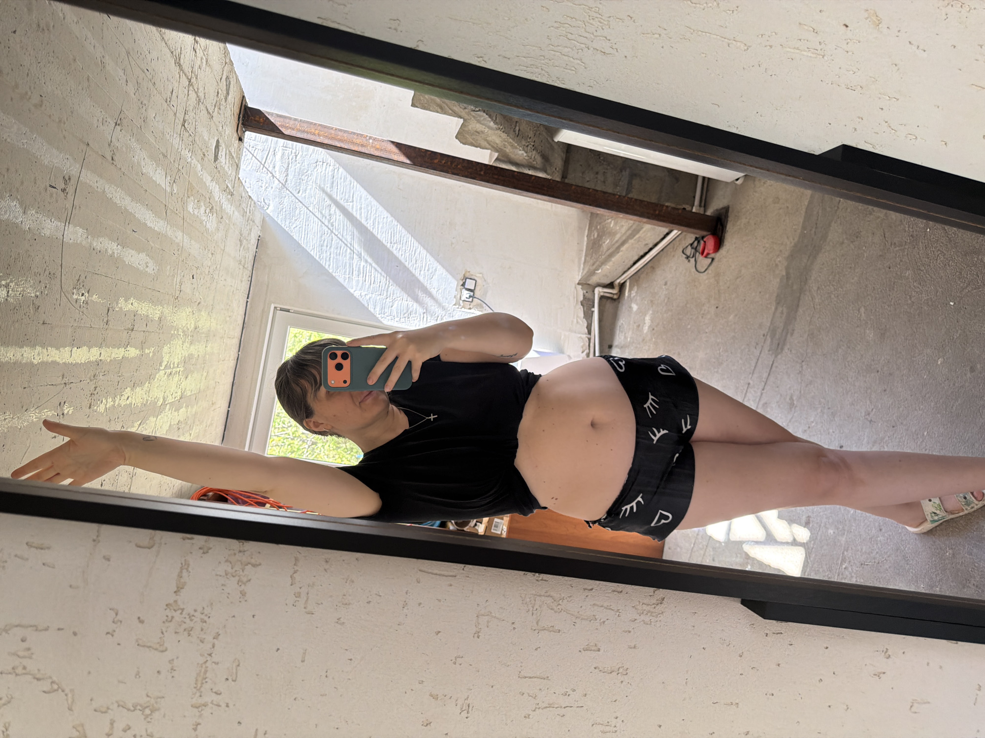 A medium shot of a person reflected in a mirror. The individual has fair skin, short hair, a black shirt, and patterned black shorts. The person is holding a smartphone, presumably taking the picture. The background appears to be a room under renovation or construction. Exposed concrete walls, support beams, and electrical outlets are visible. The floor is concrete. A glimpse of greenery can be seen through a window. The location is possibly Agoy, Russia.