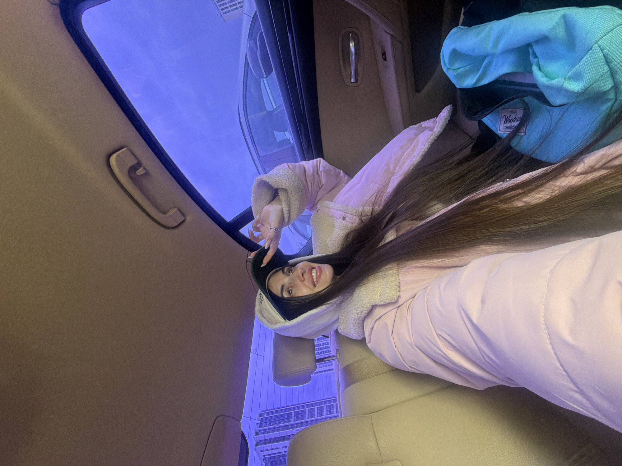 The image shows a person inside a vehicle, likely a car, with a view of a building through the sunroof. The person is wearing a light pink puffer jacket with a fur-lined hood, and a black beanie. They are smiling and making a peace sign with their hand. Another jacket, blue, is visible in the back seat. The building seen through the sunroof suggests a modern structure with multiple windows. The image seems to have been taken inside a moving vehicle. The building visible through the sunroof has distinctive architectural features, which align with the Lakhta Center in Saint Petersburg, Russia.