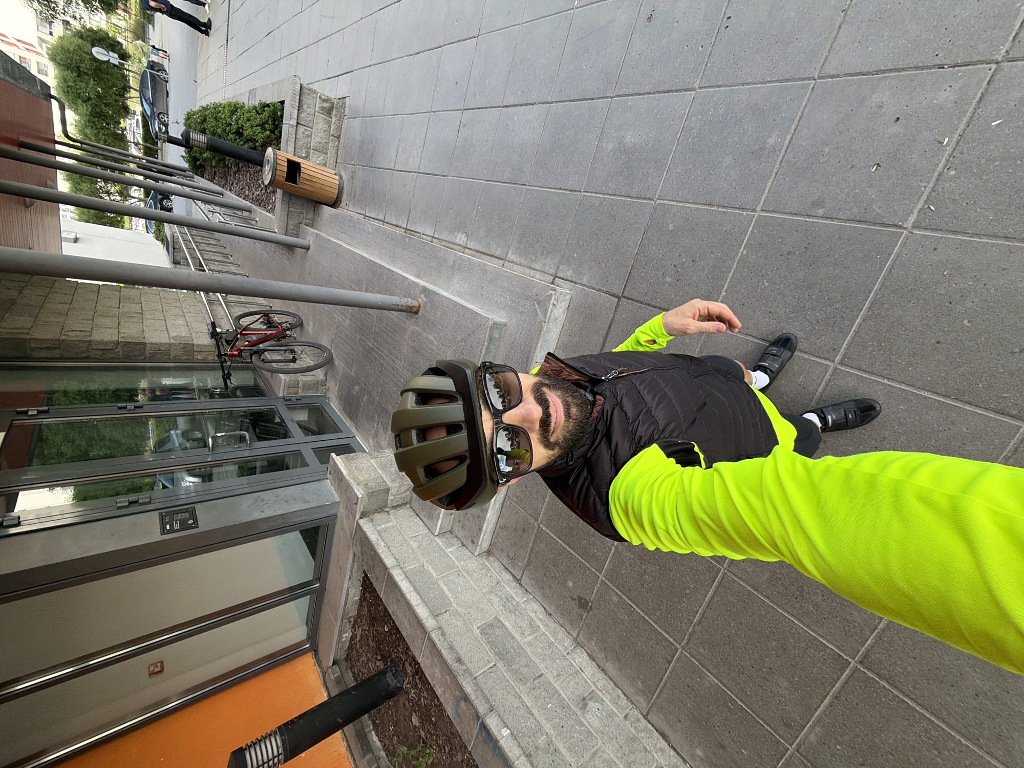 A selfie-style image, taken from a high, tilted angle, shows a man standing on an urban sidewalk. The man, the primary subject, wears an olive green cycling helmet, dark sunglasses, a black sleeveless puffer vest over a bright neon yellow long-sleeved shirt, white socks, and black shoes. He has a dark beard and appears to be holding the camera with his extended right arm.
The ground is paved with large grey square tiles. To the man's left, a red bicycle is parked next to a series of grey metal railings, likely bike racks. Behind the bicycle, a building facade with extensive glass panels and stonework is visible. Further into the scene, a wooden slatted trash can and a tall black lamp post stand near a planter box filled with green shrubs. In the background, parked cars, trees, and other urban buildings can be seen, along with a few indistinct figures.
The lighting suggests it is daytime, with soft, ambient light implying an overcast sky or early/late daylight. No legible text is visible on any signs or objects. The scene depicts a typical city street or pedestrian area. The location is Tyumen, Russia.