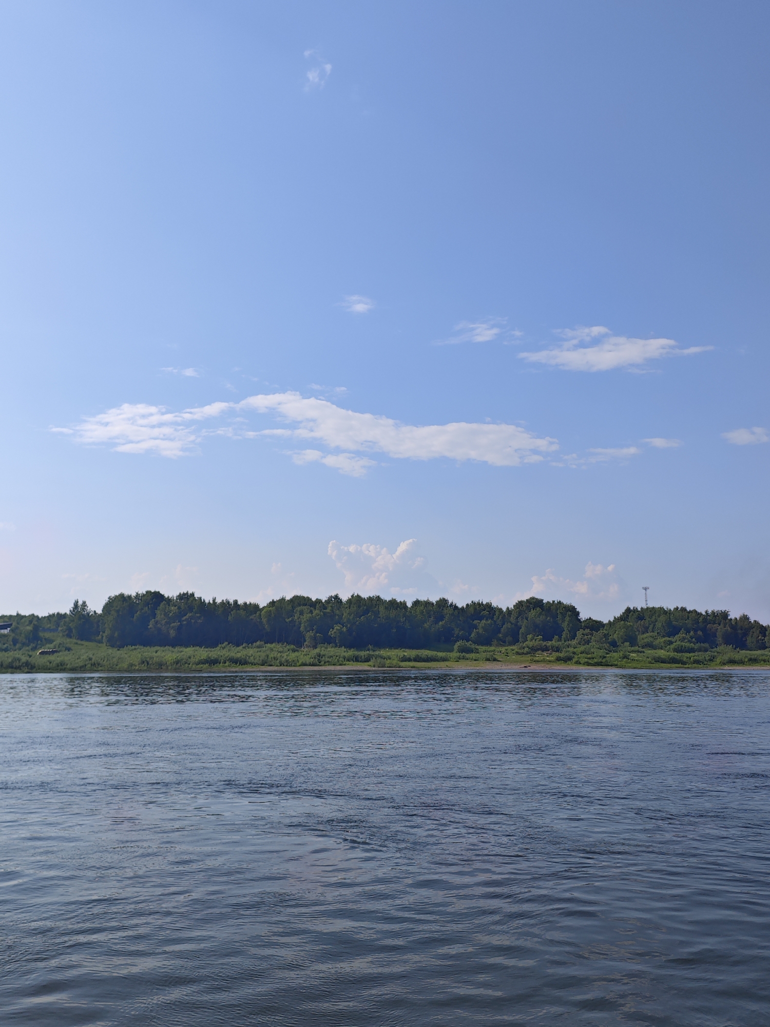 The image depicts a tranquil river scene in Верхнепашино, Russia, under a clear blue sky dotted with sparse white clouds. The foreground is dominated by the rippling surface of a wide, dark blue river, reflecting the sky. The middle ground features a lush green riverbank, densely populated with a variety of trees and vegetation. In the distance, a few buildings are visible on the left side of the bank, peeking through the foliage. On the right side of the bank, a tall, slender structure resembling a radio tower or pylon stands against the sky. The overall atmosphere is serene and natural, indicative of a peaceful day by the river.