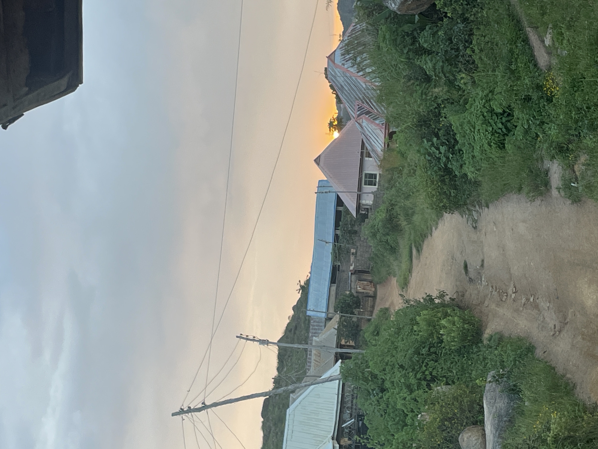 The image depicts a rural or semi-rural setting in Jos, Nigeria, characterized by a dirt path winding through overgrown vegetation. Several buildings with corrugated metal roofs are visible, suggesting a residential area. The sky is overcast with the sun setting in the distance, casting a warm glow. A telephone pole with visible wires stands prominently to the left, indicating the presence of infrastructure. The overall impression is one of a modest community nestled within a natural landscape.