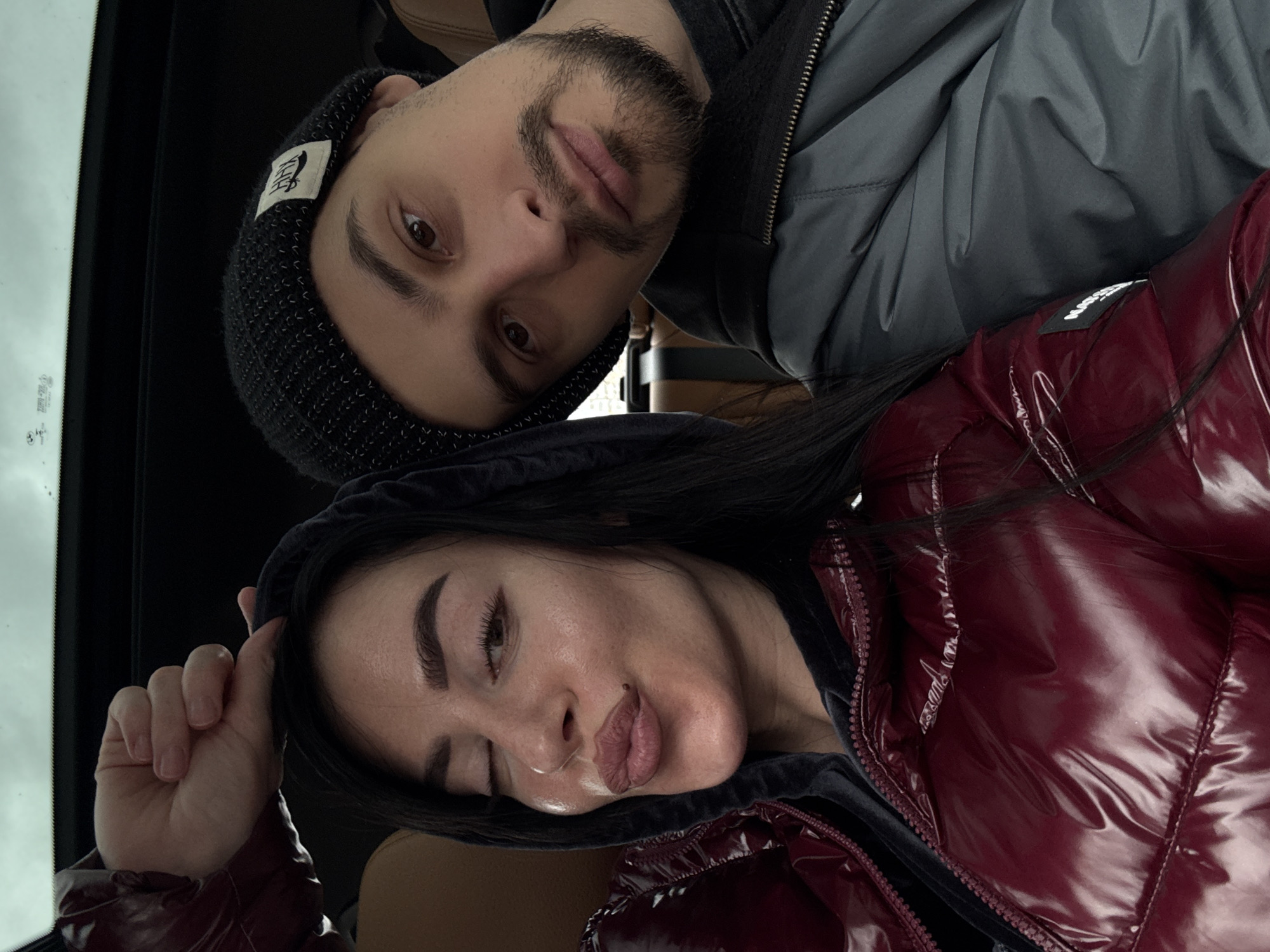 A man and a woman are in an automobile. The man is wearing a black beanie, a grey jacket, and has a beard. The woman is wearing a dark hooded jacket, and a burgundy-colored shiny puffer jacket. The car appears to be parked as a window and cloudy sky are visible in the background. The location is unknown and there is no evidence it is in Krasnoyarsk, Russia.